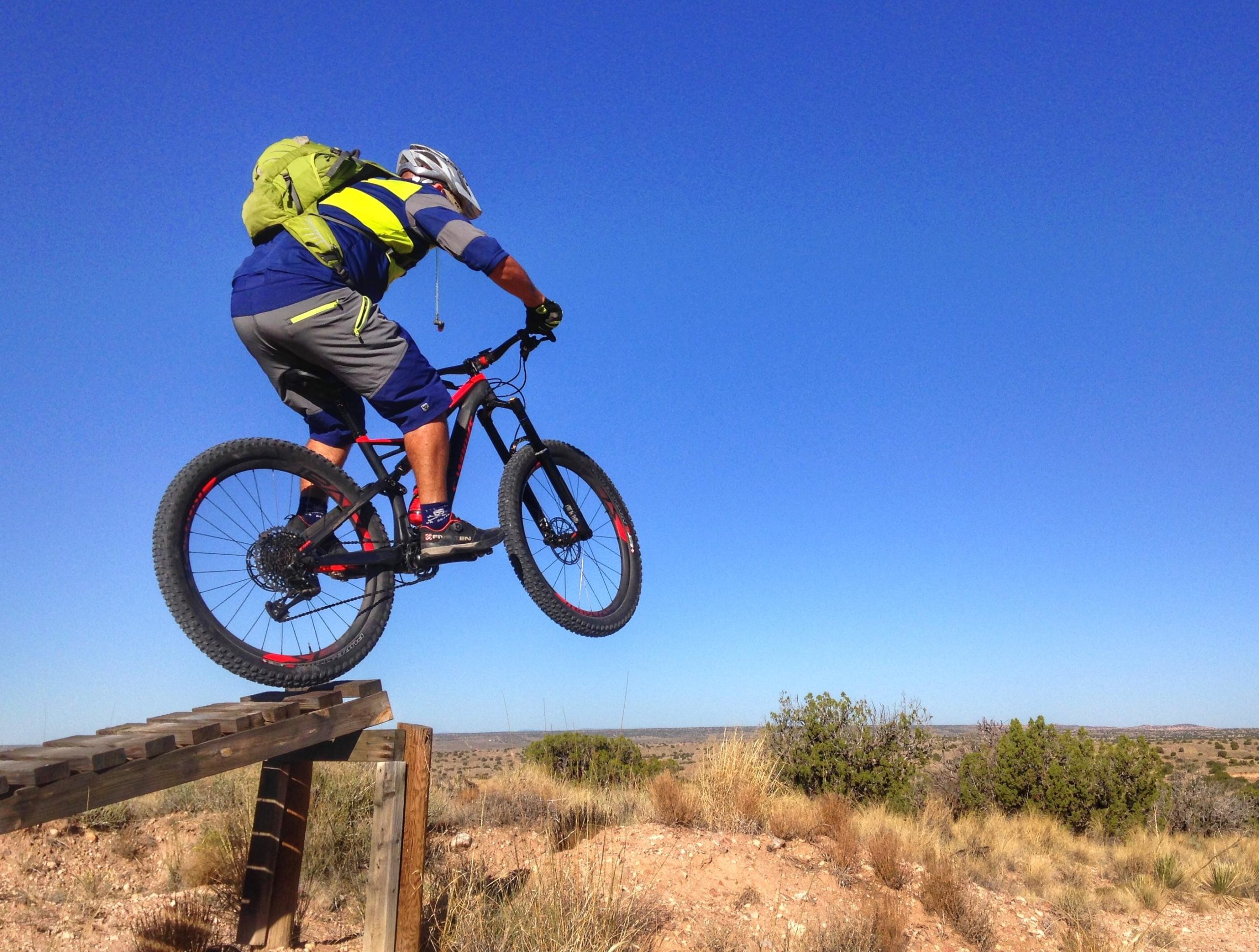 A cyclist in mid-air performing a jump on a mountain bike, wearing a helmet and a green backpack. The scene is set against a clear blue sky, with a rugged landscape of dirt and sparse vegetation in the background. Mariposa Fat Bike Trails mountain bike trail.