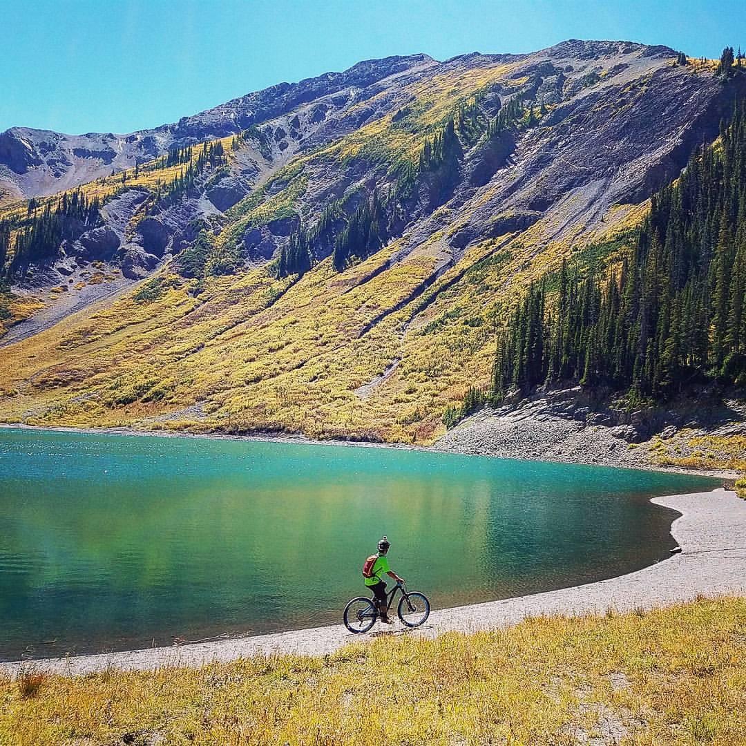 A person riding a mountain bike along the shore of a lake, surrounded by vibrant autumn foliage and rugged mountains under a clear blue sky. Trail 401 mountain bike trail.