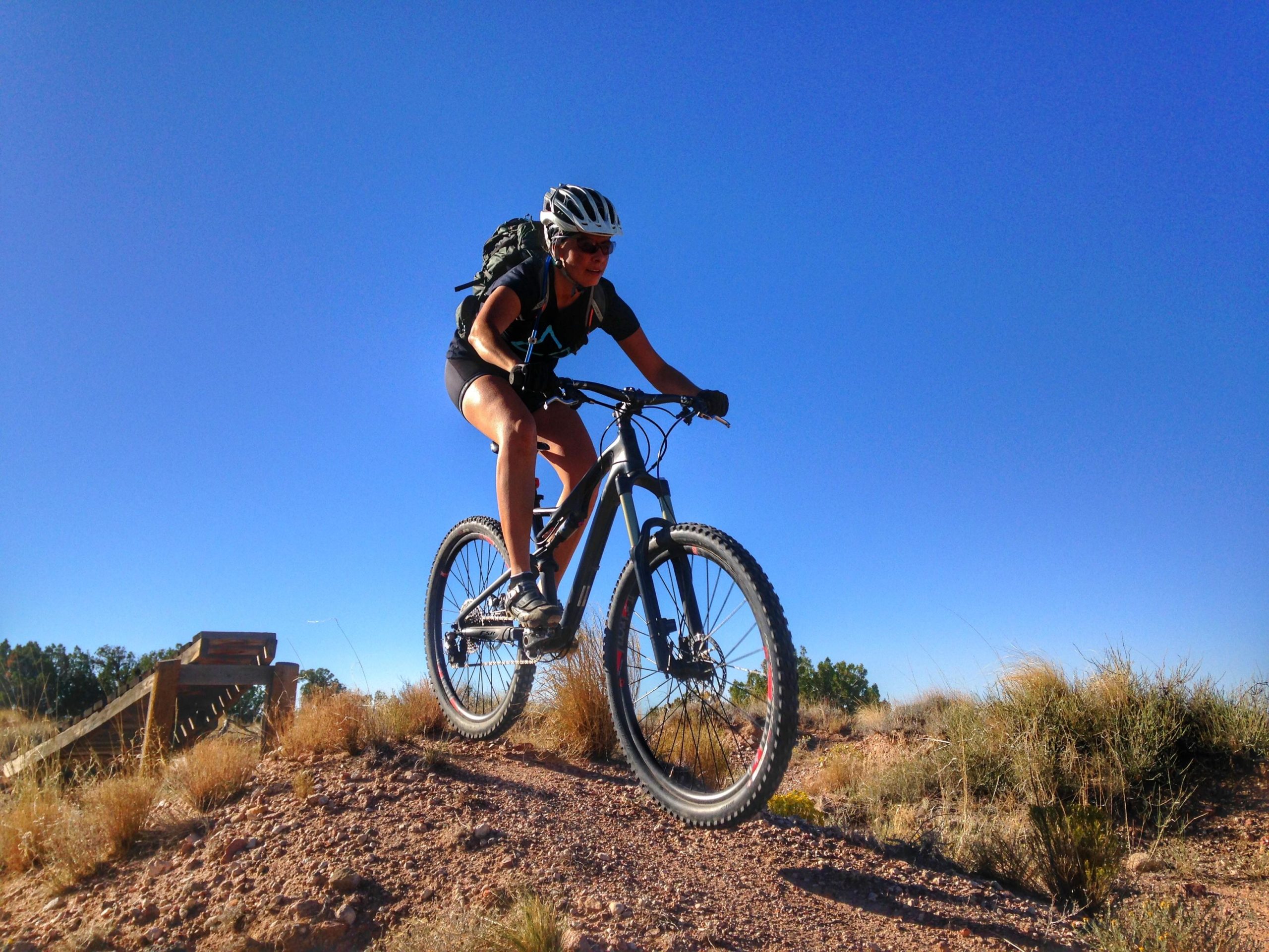 A person riding a mountain bike on a dirt trail, leaning forward with focused determination. The rider is wearing a helmet and sunglasses, and is surrounded by sparse vegetation under a clear blue sky. A wooden ramp is visible in the background. Mariposa Fat Bike Trails mountain bike trail.