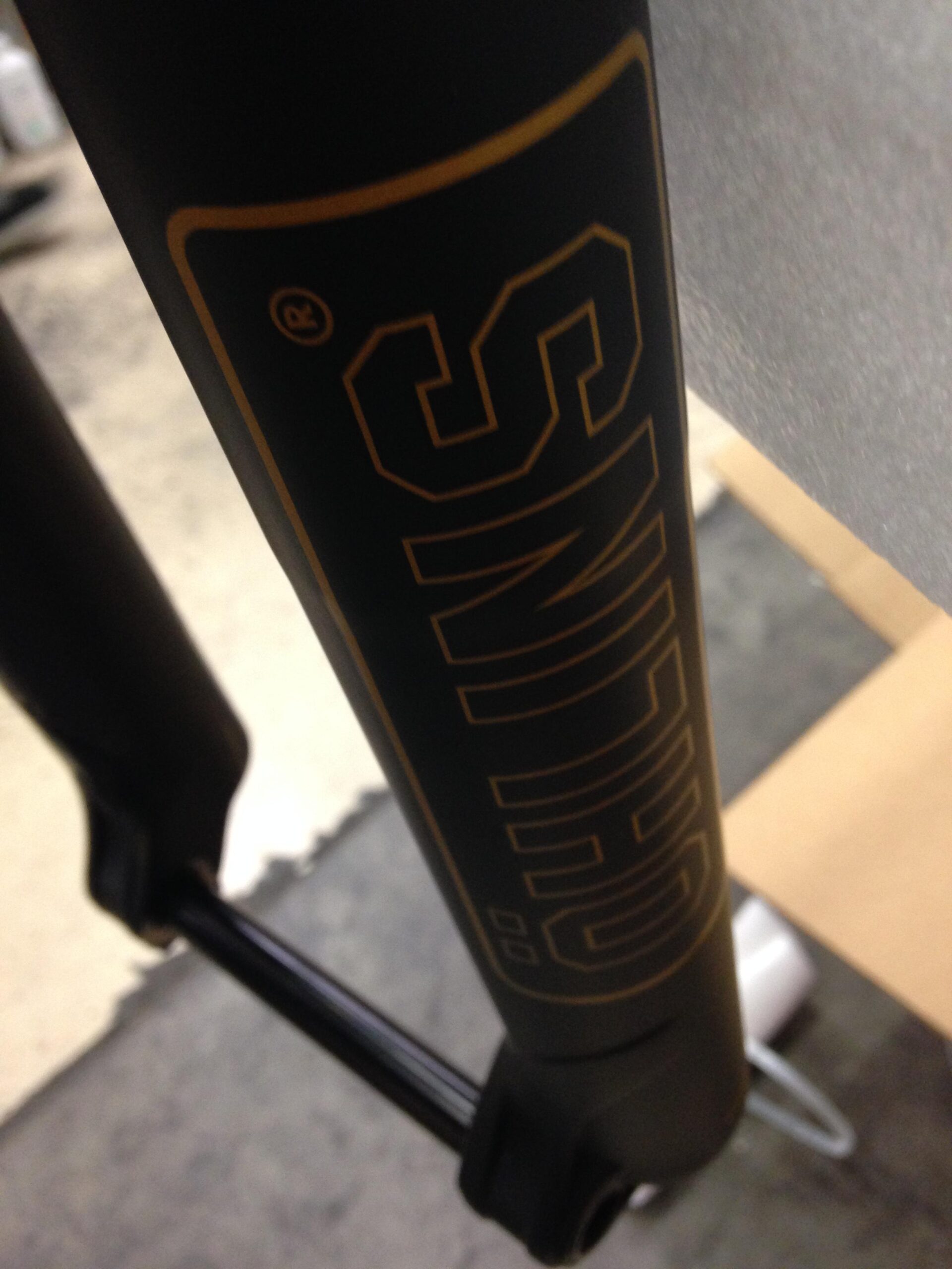 Close-up view of a black bicycle fork leg featuring a prominent gold logo. The image showcases the branding on the fork, with part of the leg and a black axle also visible in the composition. The background is blurred, emphasizing the details of the fork and its design.