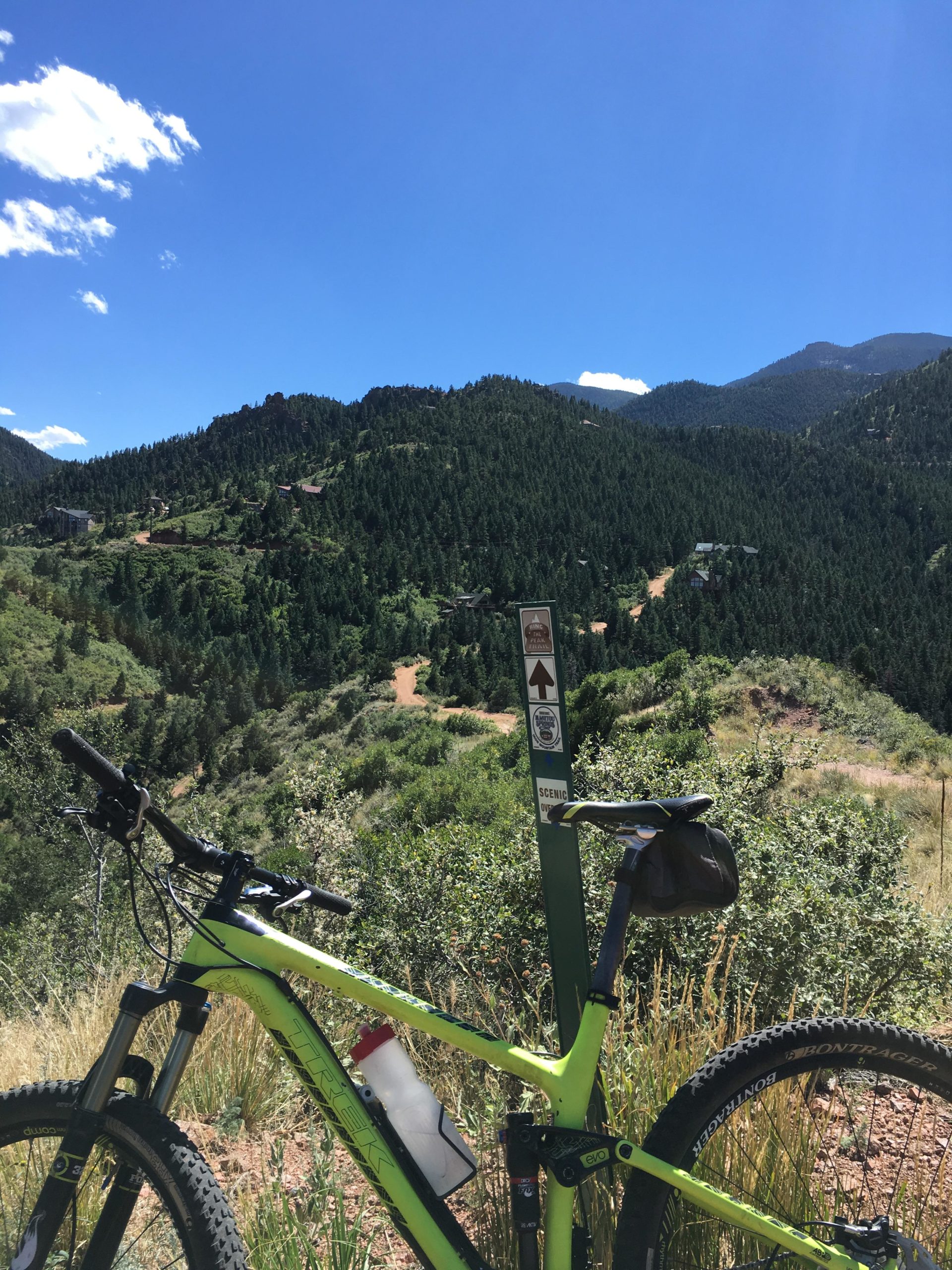 A mountain bike stands in the foreground, with a green frame and a water bottle attached, positioned next to a trail sign in a lush forested area. In the background, rolling hills and mountains rise under a bright blue sky with scattered clouds. The scenery suggests a recreational outdoor setting ideal for biking and hiking. Paul Intemann Memorial Trail mountain bike trail.