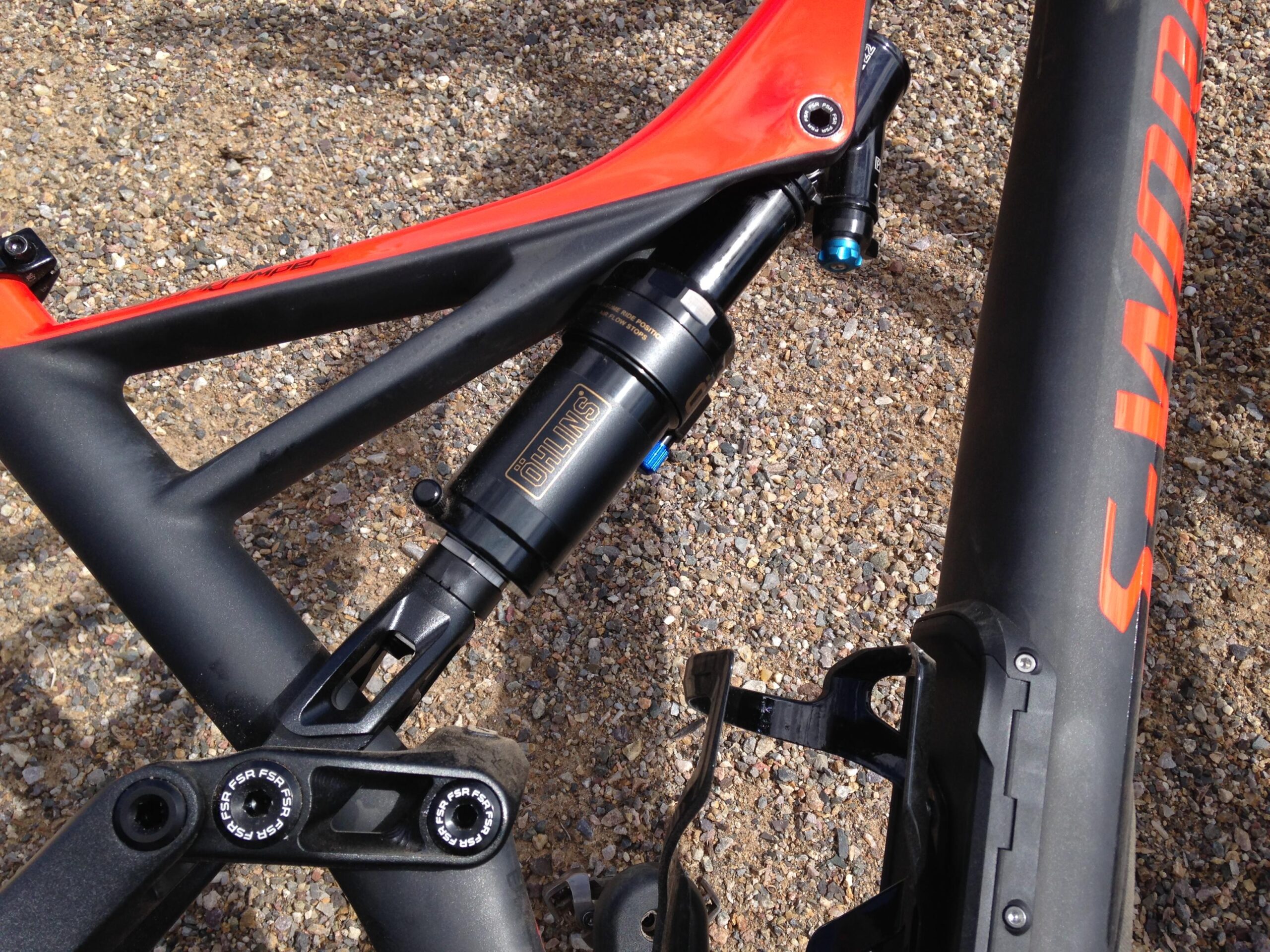 Close-up view of a mountain bike frame featuring a black and red color scheme. The image highlights a shock absorber marked with the brand "Öhlins" mounted within the frame's rear triangle. The bike's carbon fiber finish is visible, along with adjustments for the shock and a water bottle holder on the side. The background consists of a gravel surface.