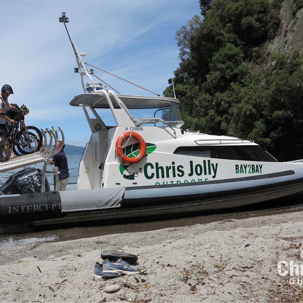 A boat named "Chris Jolly Outdoors" is anchored on a sandy shore. Two individuals are loading bicycles onto a bike rack on the boat while a third person watches. Nearby, a pair of shoes is resting on the sand, and the surrounding area features lush greenery and a calm body of water. Great Lake Trails mountain bike trail.