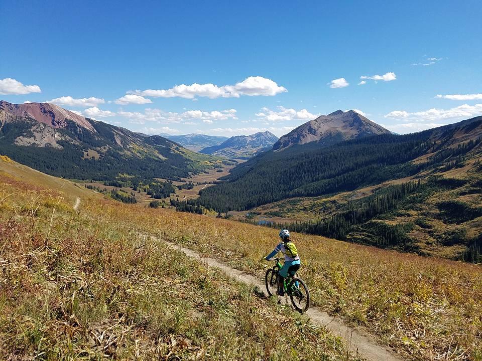 A mountain biker rides along a dirt trail on a hillside, surrounded by vibrant autumn foliage and expansive mountain views under a clear blue sky with scattered clouds. Trail 401 mountain bike trail.
