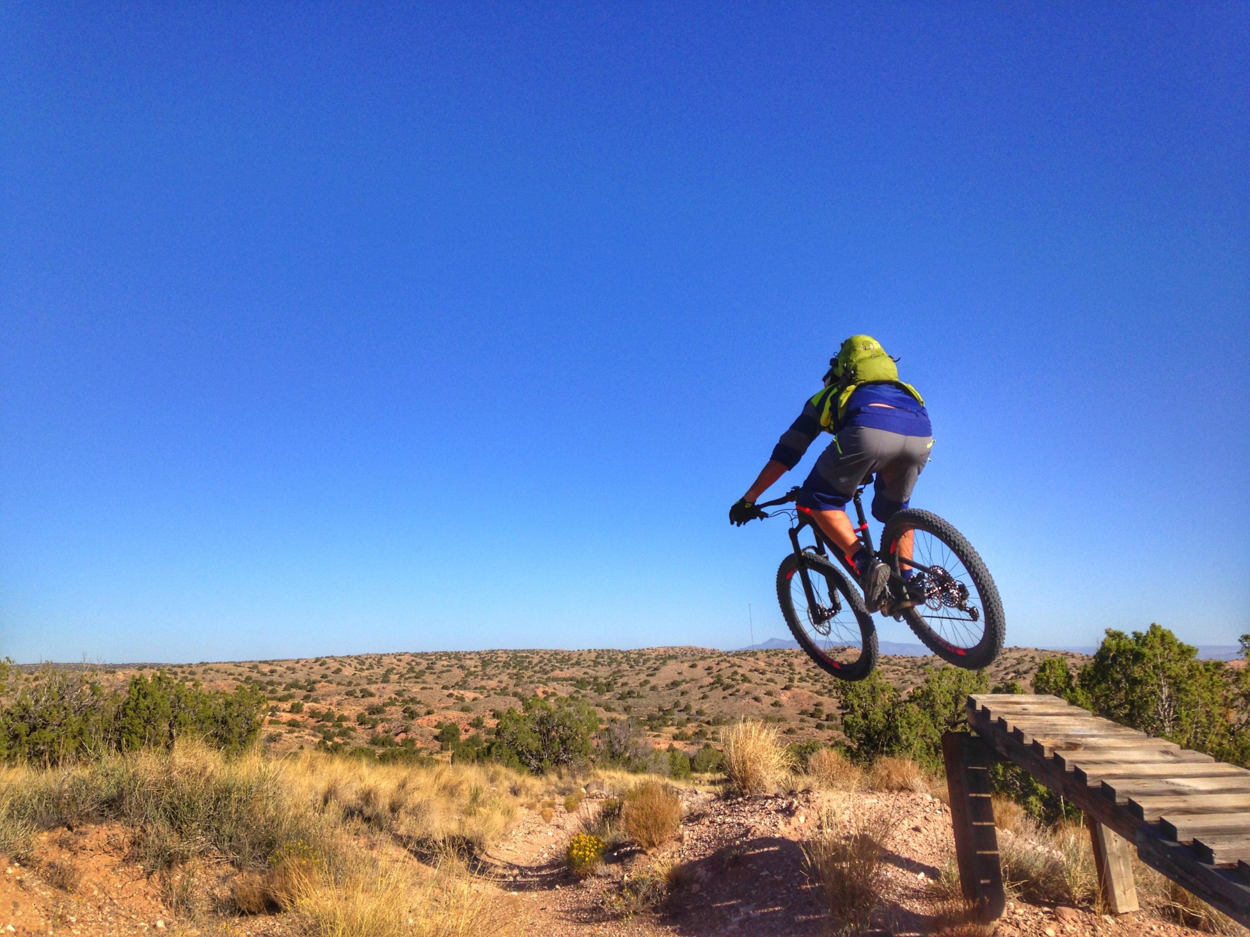 A mountain biker jumps off a wooden ramp against a clear blue sky, with a landscape of shrubs and hills in the background. The biker is wearing a helmet and sports gear, showcasing an action-filled moment in outdoor cycling. Mariposa Fat Bike Trails mountain bike trail.