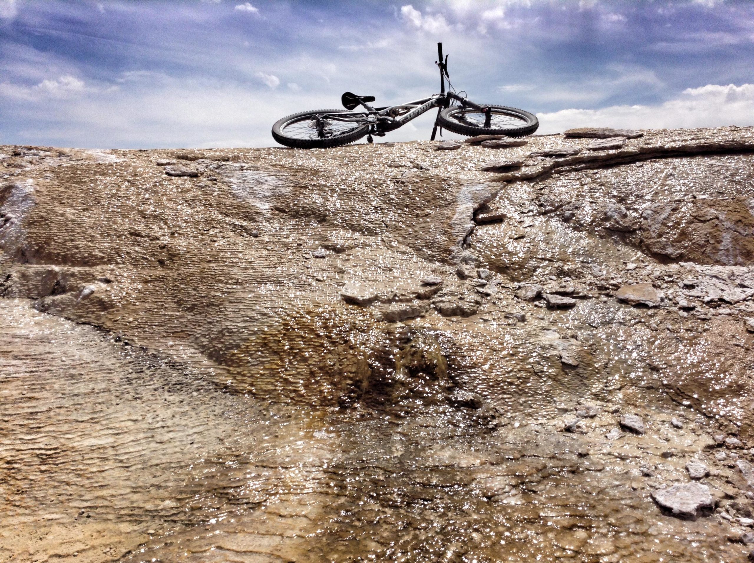A mountain bike resting on a rocky surface with shimmering water, under a bright blue sky with scattered clouds. White Ridge Bike Trails mountain bike trail.