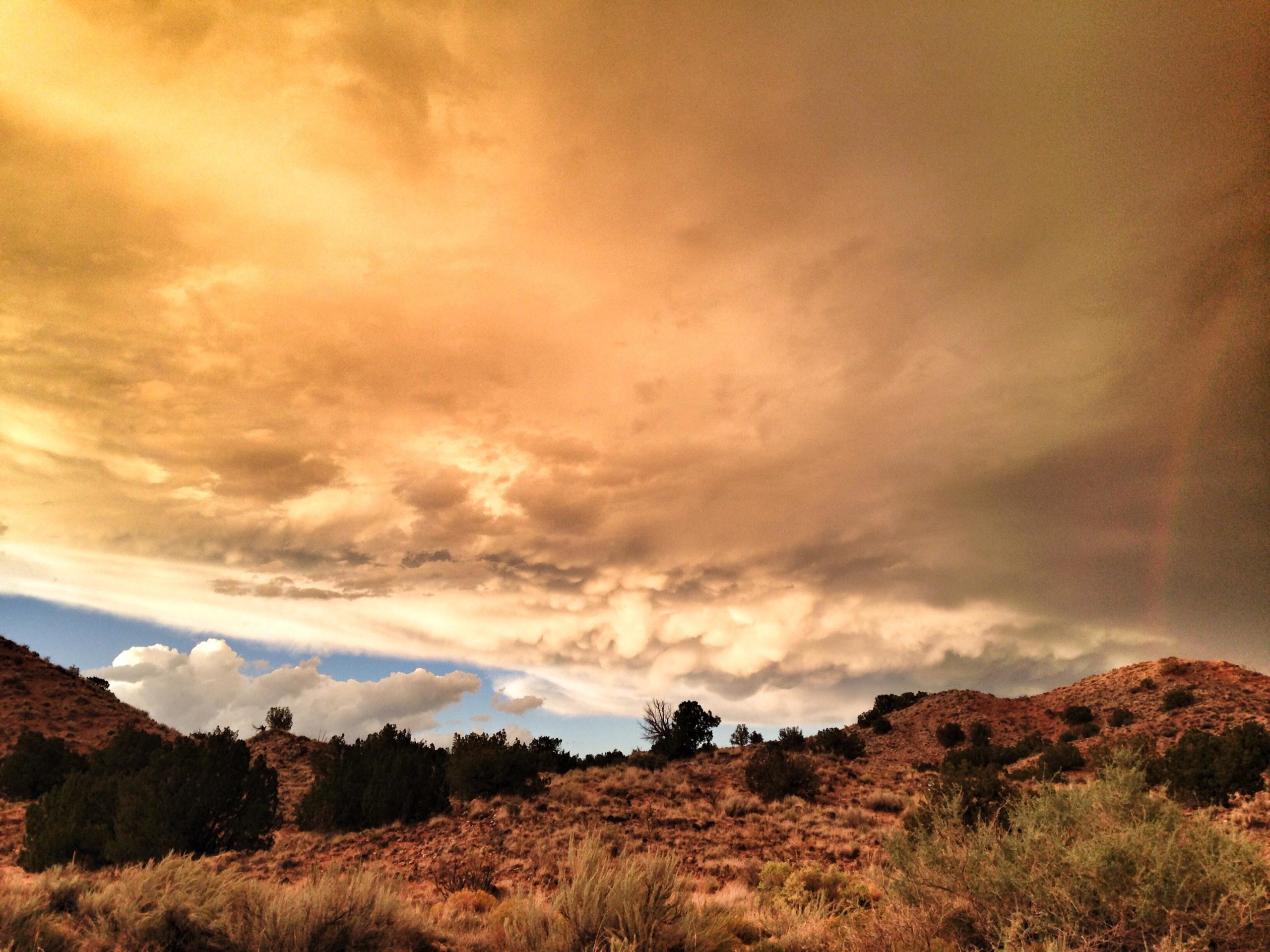 Wide landscape featuring rolling hills with sparse vegetation, under a dramatic sky filled with layers of clouds illuminated in warm, orange and golden hues. The scene captures the interplay of light and shadow, suggesting an impending storm or sunset. Mariposa Fat Bike Trails mountain bike trail.