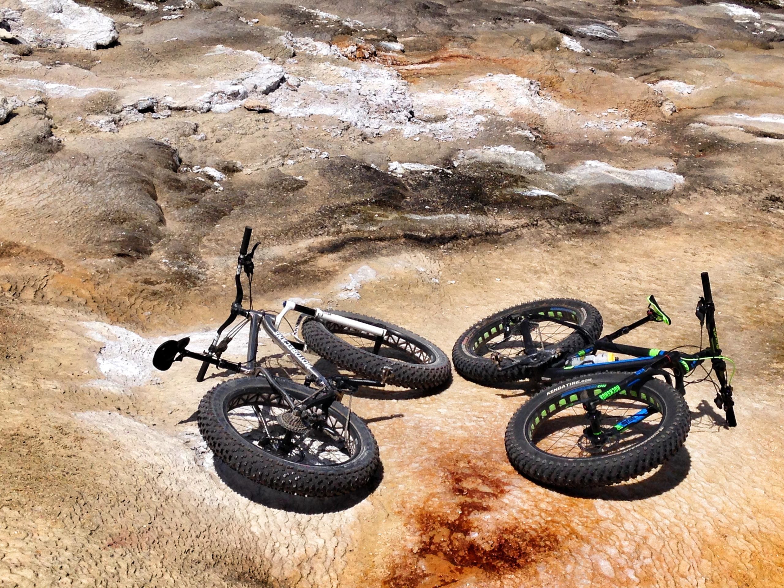 Two mountain bikes with large tires are lying on rugged, uneven terrain featuring a mix of earthy tones and patches of white mineral deposits. The scene suggests an outdoor adventure, possibly in a natural park or hilly area. White Ridge Bike Trails mountain bike trail.