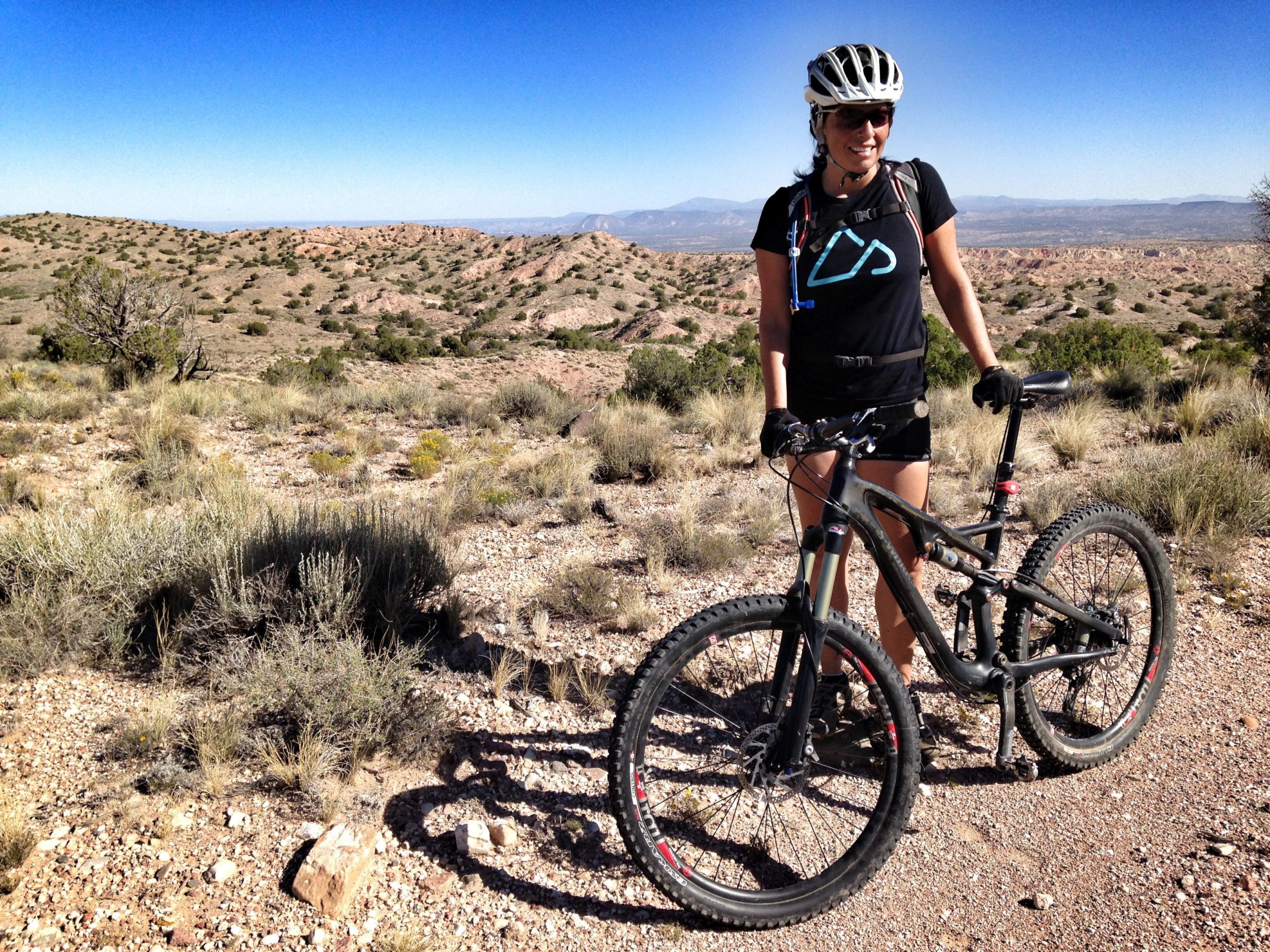 A cyclist stands next to a mountain bike on a rocky trail, surrounded by a desert landscape with rolling hills and sparse vegetation. The sun is shining in a clear blue sky, and the cyclist is dressed in a black shirt and shorts, wearing a helmet and gloves, and smiling. Mariposa Fat Bike Trails mountain bike trail.