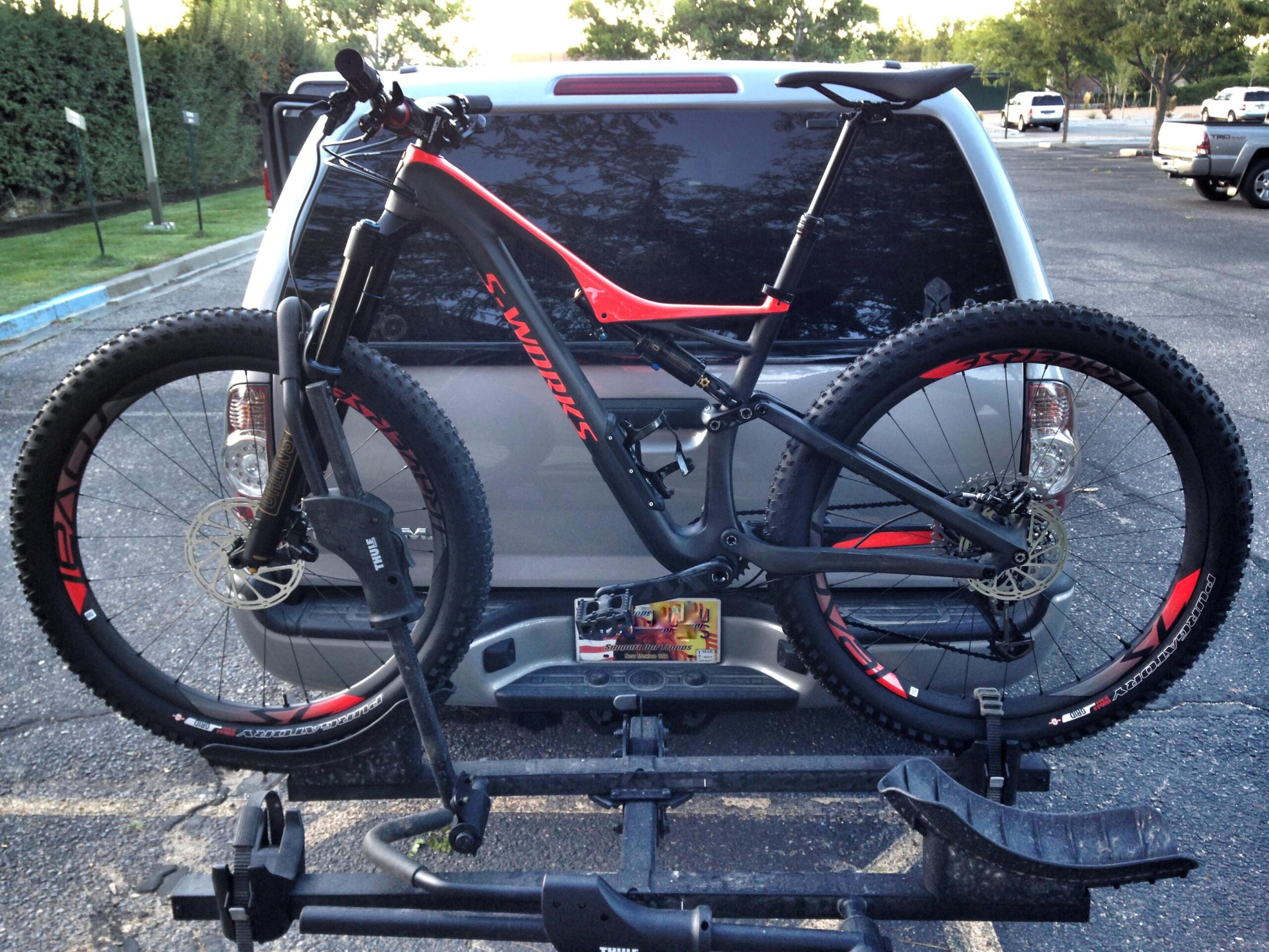 Specialized Stumpjumper FSR 6Fattie: A high-performance mountain bike mounted on a bike rack attached to the rear of a gray SUV, surrounded by parking spaces and greenery in the background. The bike features a sleek black and red design, wide tires, and a suspension fork.
