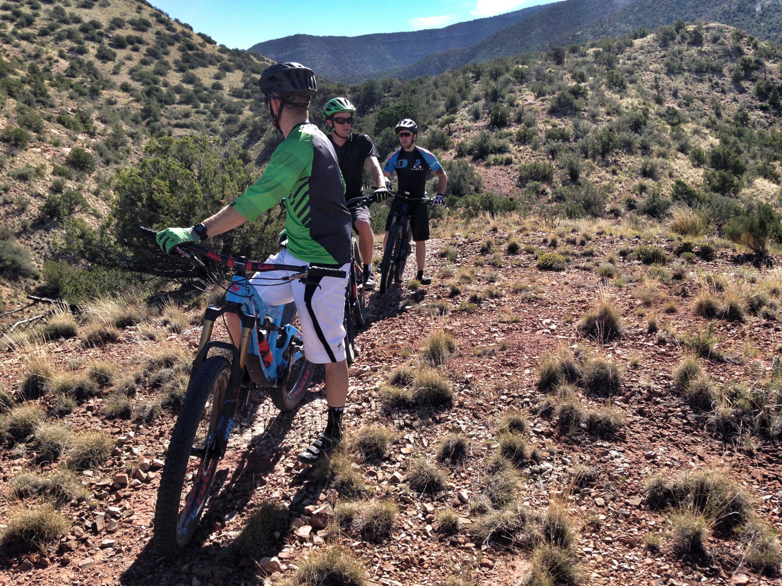 Three mountain bikers are standing on a rocky trail surrounded by hills and sparse vegetation. The riders are dressed in athletic gear and helmets, with two bikes positioned next to them. The scene captures a beautiful outdoor landscape under a clear blue sky. Placitas Trails mountain bike trail.