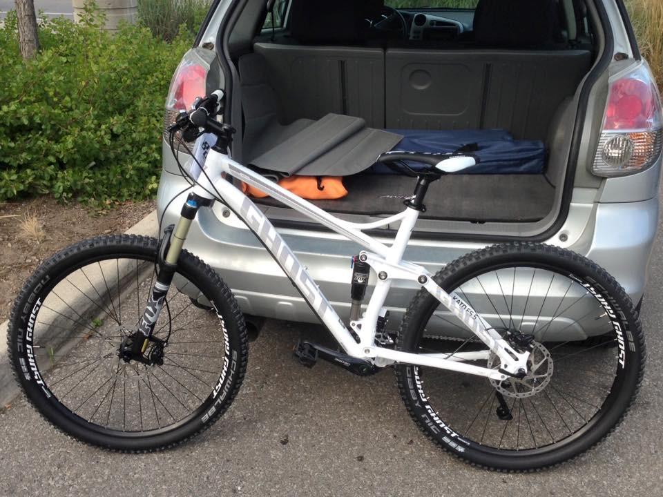 Ghost Kato FS 5: A mountain bike with a white frame is parked next to the open rear hatch of a silver car. The bike features large, knobby tires and front suspension, with gear and a folded blanket visible in the car's interior. In the background, there is a patch of greenery.