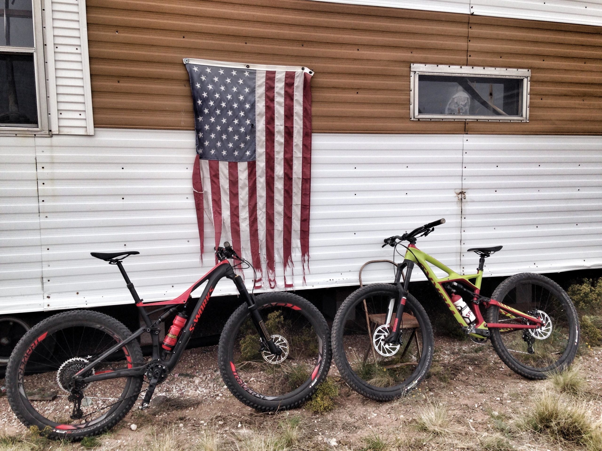Two mountain bikes, one black and red, the other green and red, are parked on the ground in front of a trailer. An American flag hangs on the side of the trailer, which features a mix of brown and white siding. A small window is visible above the bikes. The surrounding area has dry grass and sparse vegetation. Mariposa Fat Bike Trails mountain bike trail.