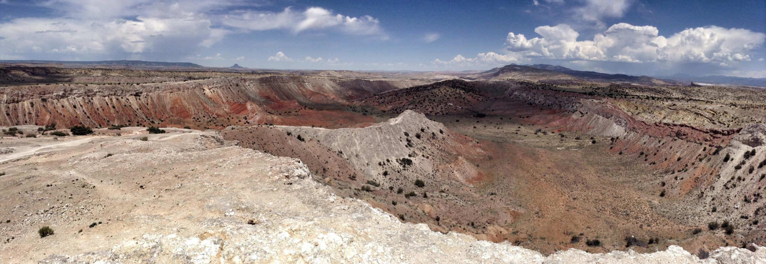 A panoramic view of a colorful desert landscape featuring layered rock formations, rolling hills, and open sky with scattered clouds. The terrain exhibits shades of red, orange, and gray, showcasing the natural erosion patterns of the earth. White Ridge Bike Trails mountain bike trail.
