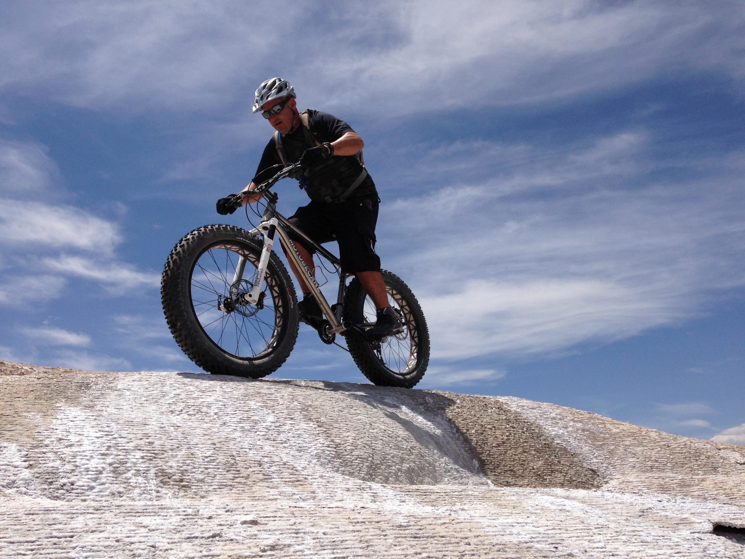 A cyclist riding a fat tire bike on a rocky surface, navigating a small incline under a blue sky with wispy clouds. The cyclist is wearing a helmet and sunglasses, appearing focused and in motion. White Ridge Bike Trails mountain bike trail.
