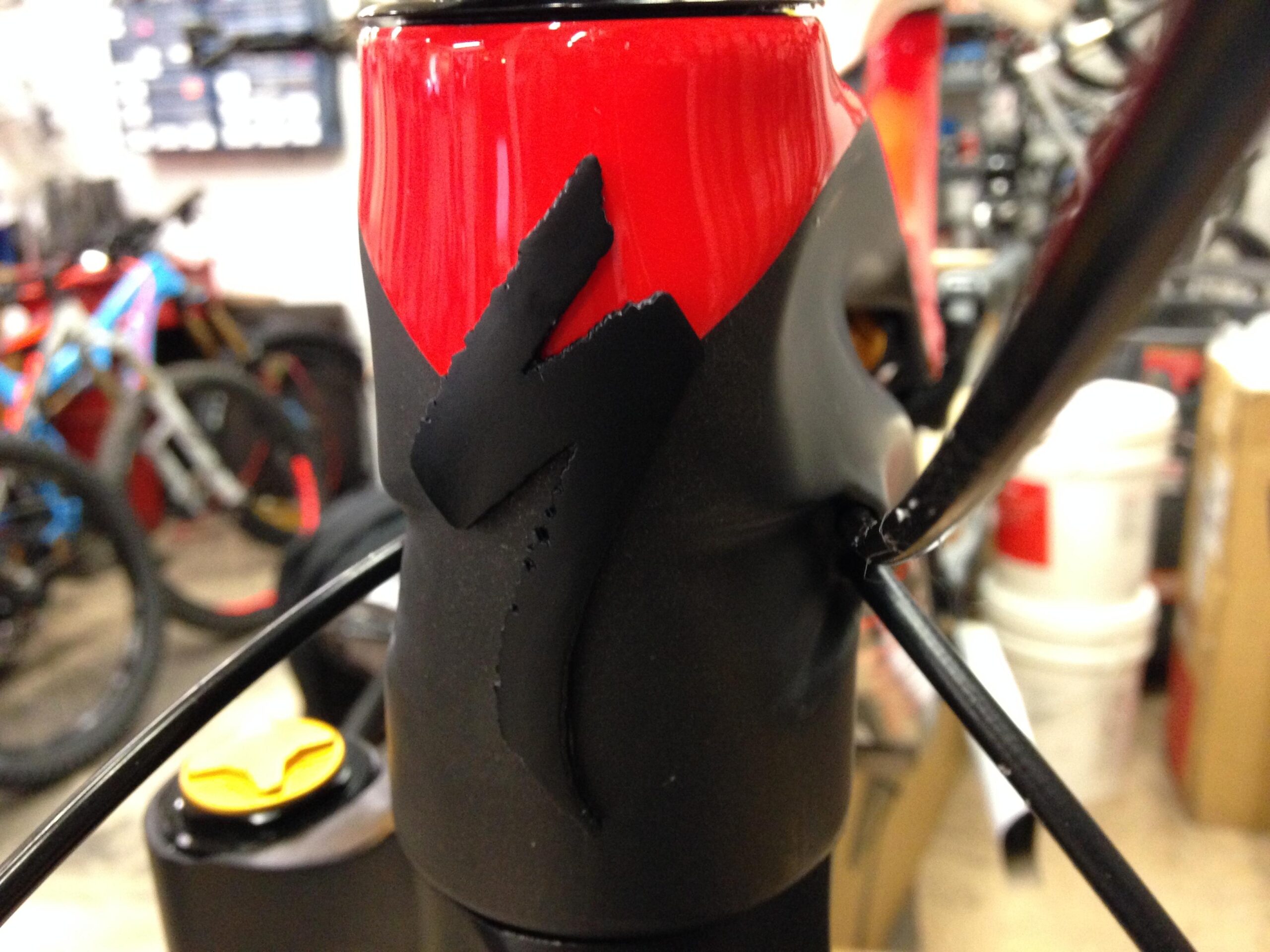 Image of a close-up view of a bicycle frame featuring a red and black design with the Specialized logo prominently displayed. The background shows a workshop setting with other bicycles and storage items visible.