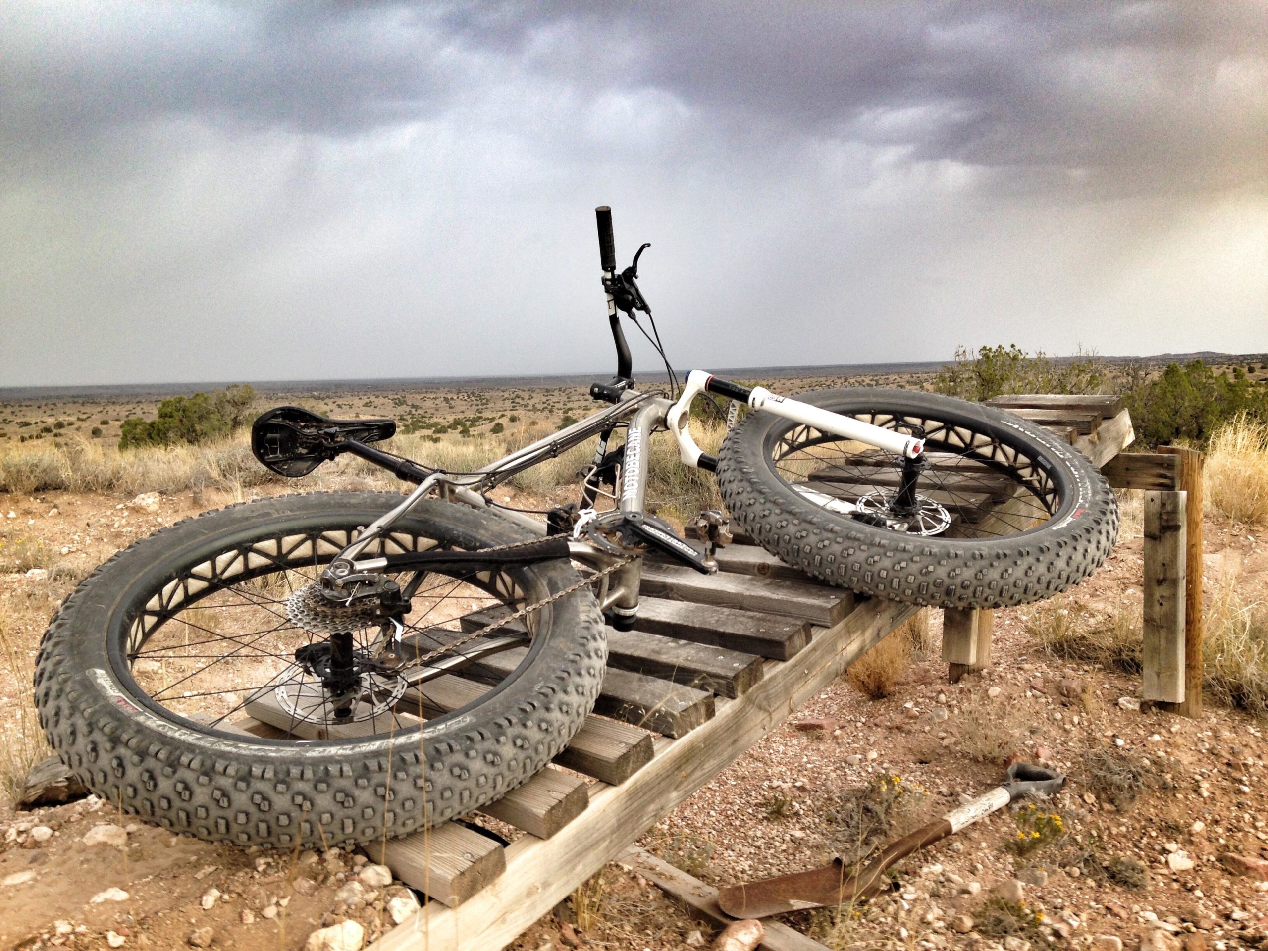 A fat tire bicycle is resting on a wooden bridge overlooking a vast, arid landscape under a cloudy sky. The ground is rocky and covered with sparse vegetation, while the horizon stretches out in the distance. Mariposa Fat Bike Trails mountain bike trail.