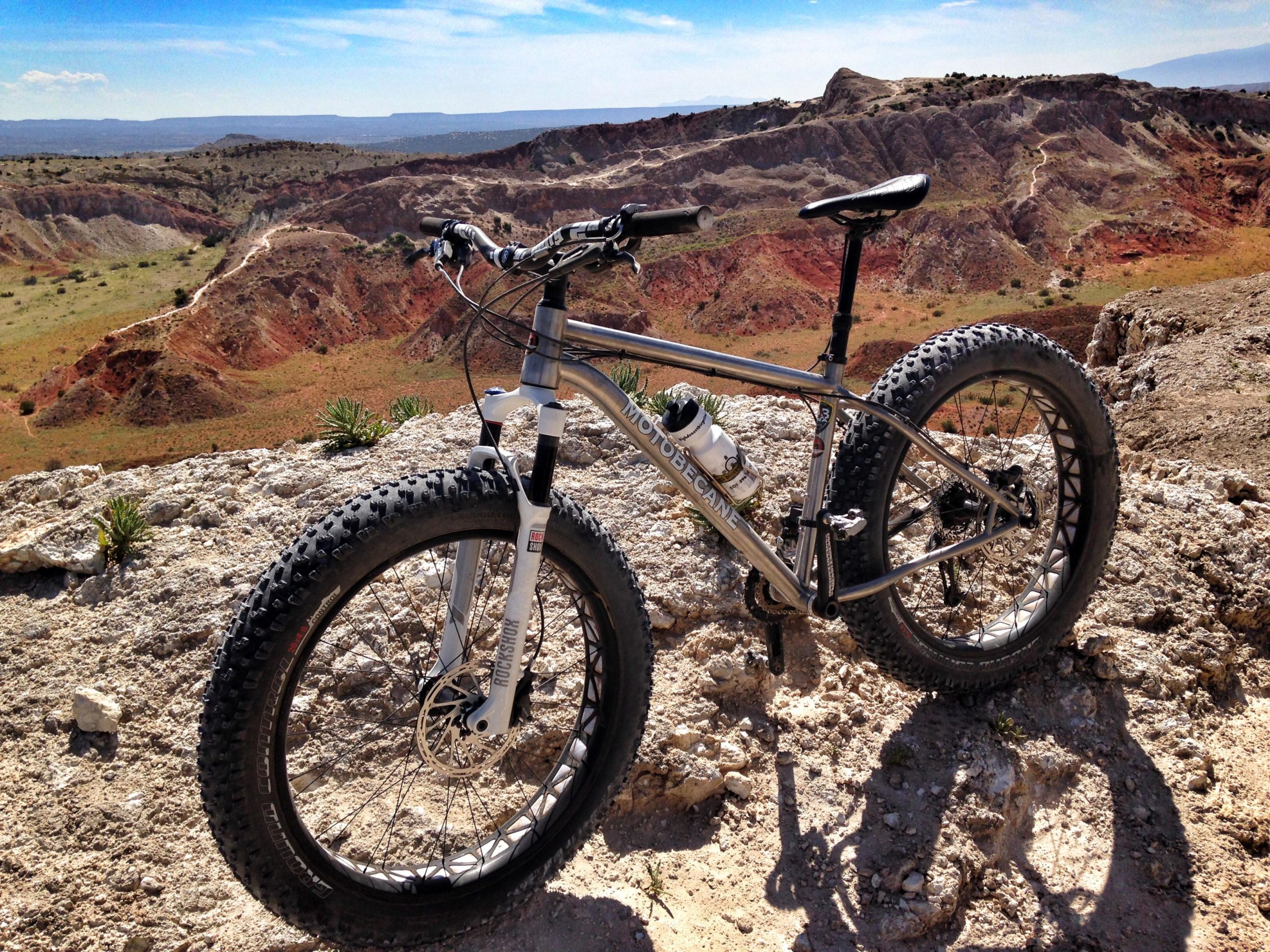 A fat tire mountain bike is positioned on rocky terrain, overlooking a rugged landscape with colorful hills and valleys under a clear blue sky. The bike features a sleek silver frame and wide tires, suggesting it is suitable for off-road riding. White Ridge Bike Trails mountain bike trail.