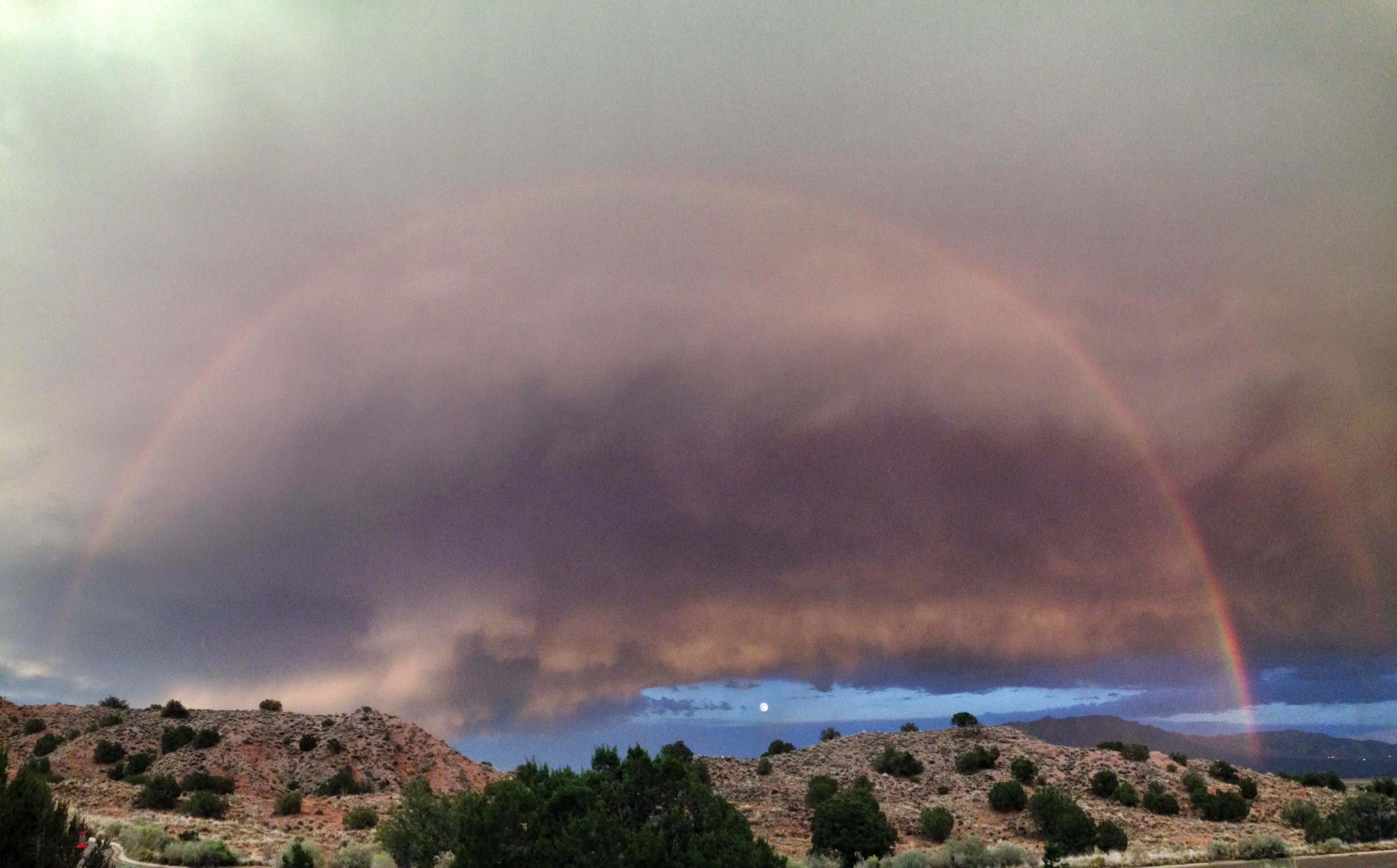 A scenic landscape featuring a vibrant rainbow arching over a rugged terrain, with dark, dramatic clouds above. The sky displays shades of gray and hints of blue, while a soft light illuminates the horizon, creating a serene and atmospheric scene. Sparse vegetation and hills are visible in the foreground, complementing the expansive sky. Mariposa Fat Bike Trails mountain bike trail.