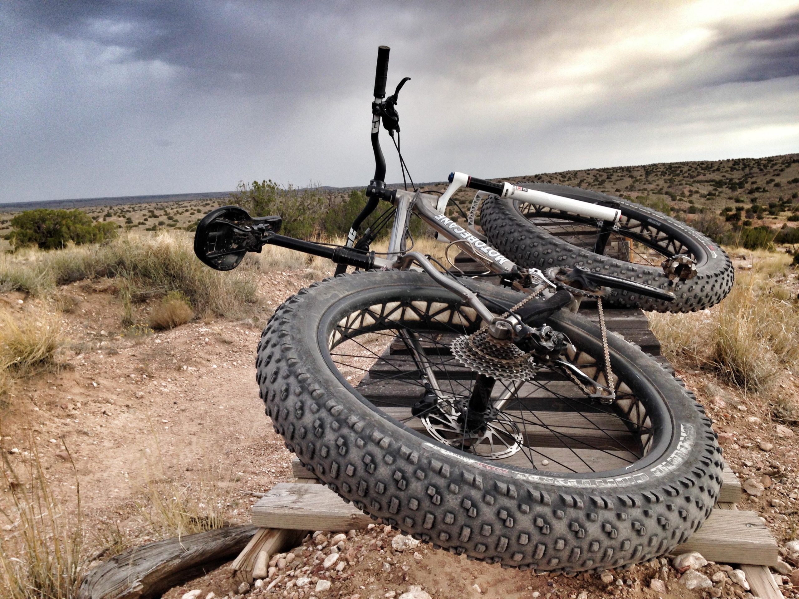 A close-up view of a mountain bike lying on its side on a dirt trail, with a rugged landscape in the background. The bike features large, textured tires and a partially visible gear system. The sky is overcast, suggesting a possibly rainy atmosphere, while surrounding grass and shrubs indicate a natural outdoor setting. Mariposa Fat Bike Trails mountain bike trail.