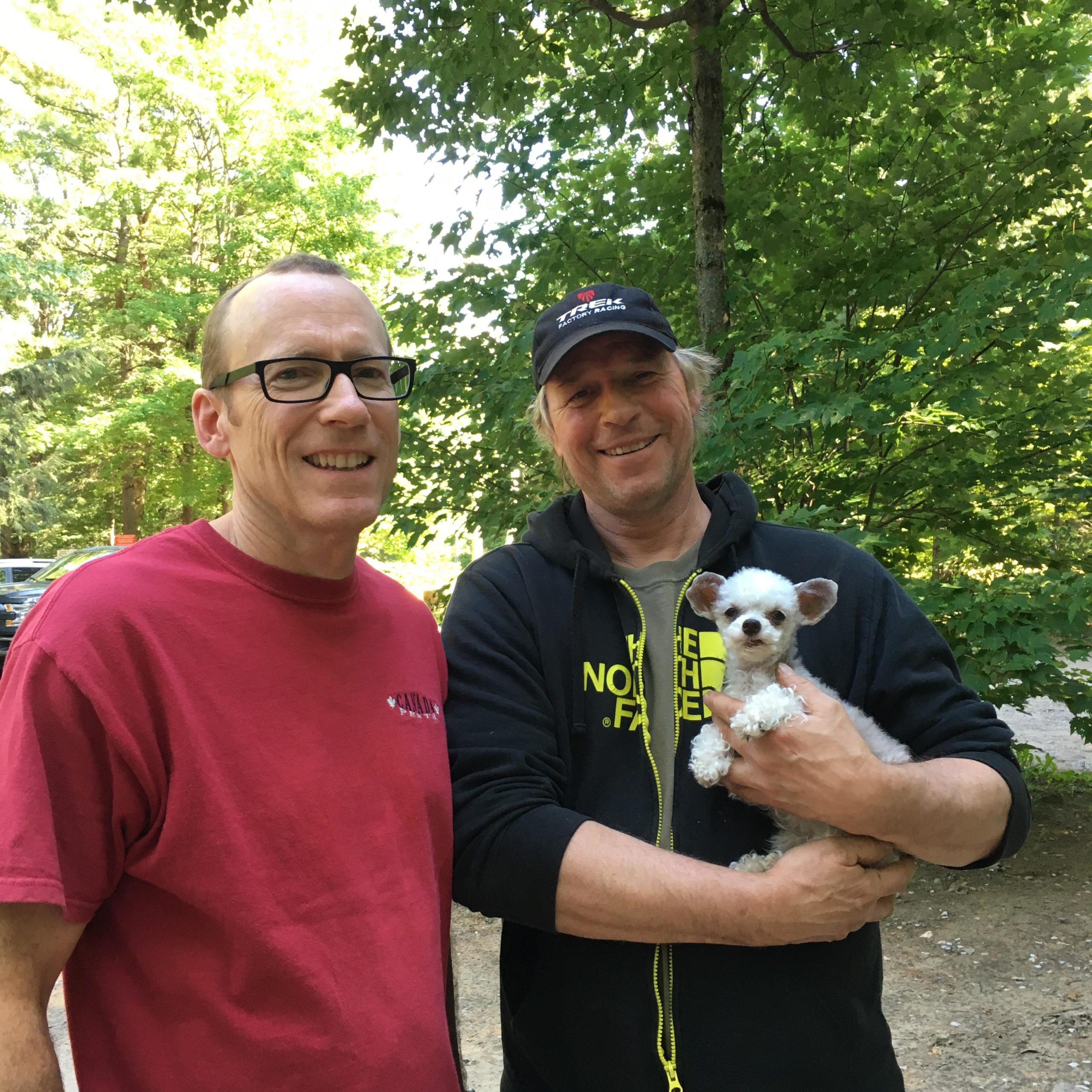 Two men stand together in a wooded area, smiling at the camera. One man, wearing glasses and a red shirt, is on the left, while the other, in a black jacket and cap, holds a small white dog. Bright green trees provide a natural backdrop. Buckwallow mountain bike trail.