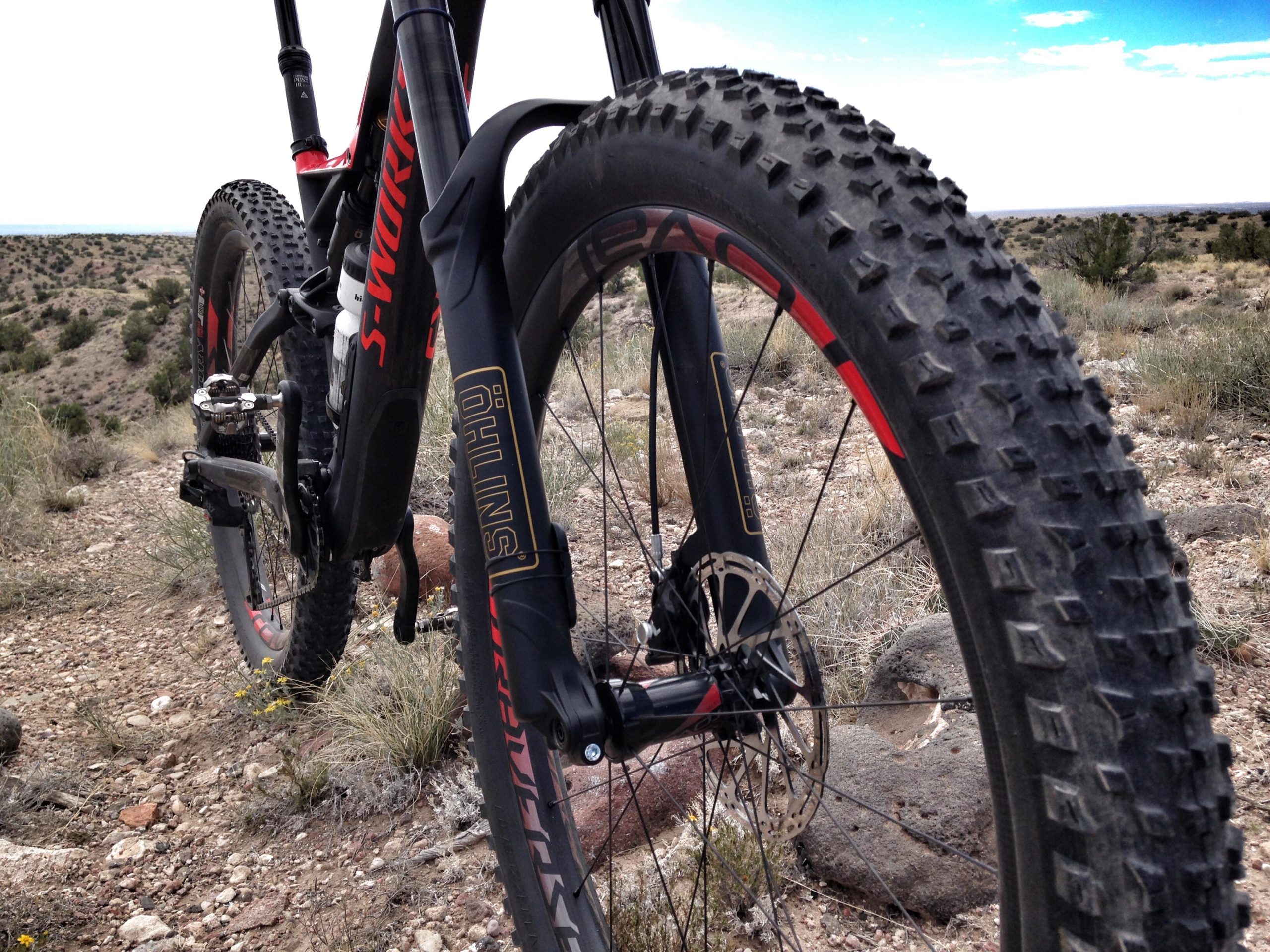 Close-up view of a mountain bike's front wheel and suspension system, set against a rugged outdoor landscape featuring sparse vegetation and rocky terrain. The bike has a prominent brand logo and a distinctive tread pattern on the tire, indicating suitability for off-road riding. Mariposa Fat Bike Trails mountain bike trail.