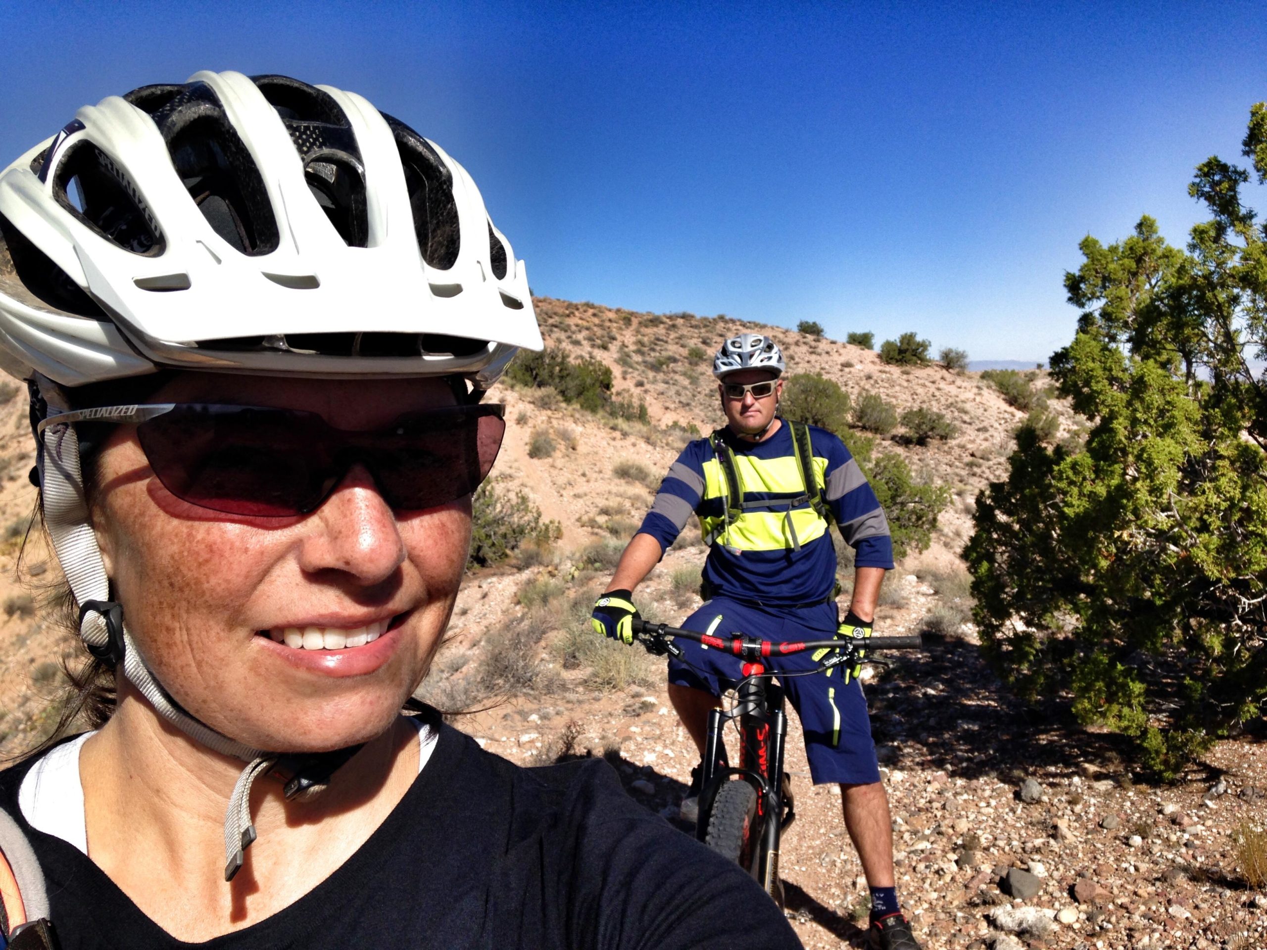 A woman in a bike helmet and sunglasses poses for a selfie on a mountain biking trail, while a man in a high-visibility outfit stands next to his bike in the background. The landscape features rocky terrain and sparse vegetation under a clear blue sky. Mariposa Fat Bike Trails mountain bike trail.