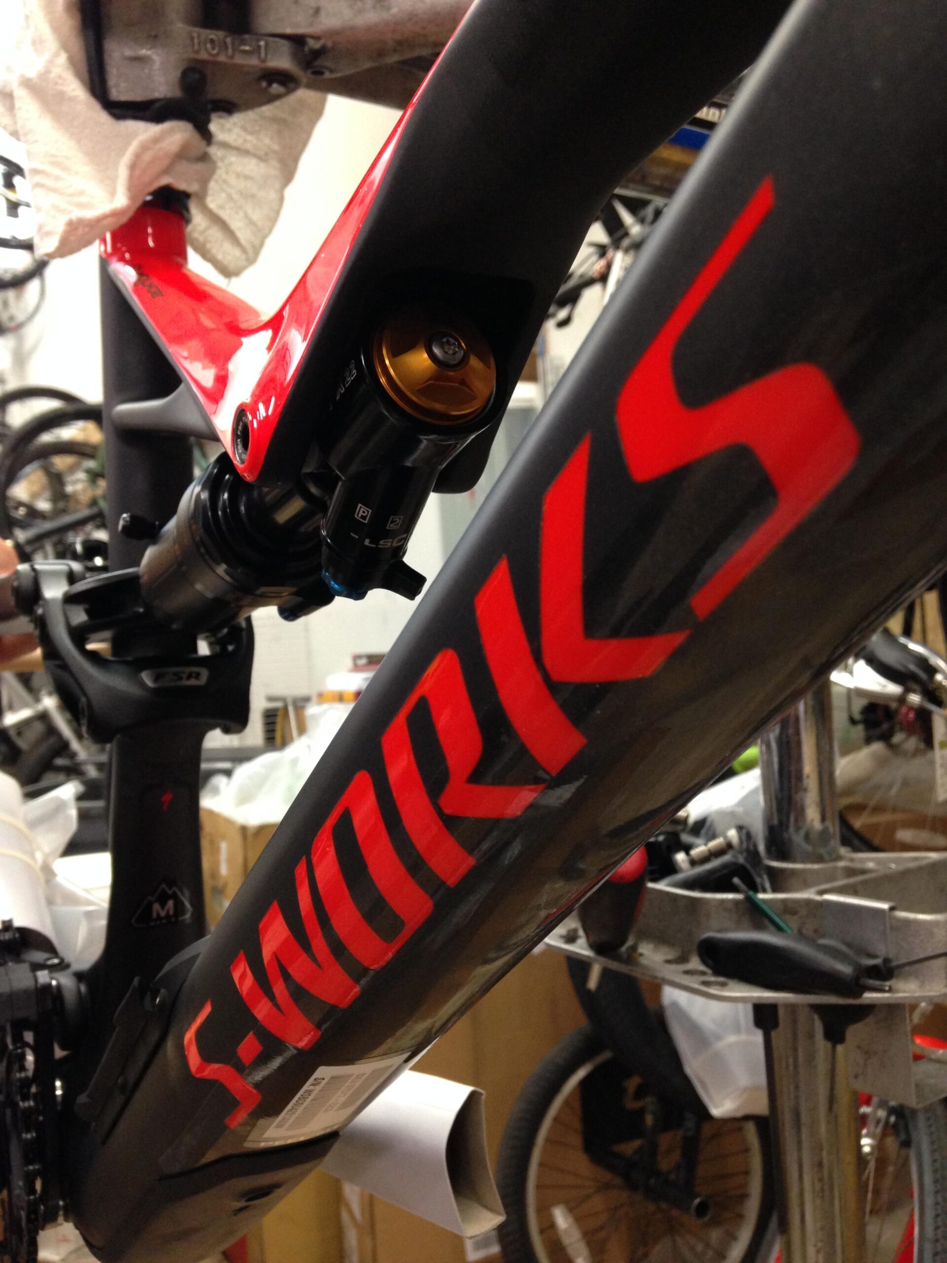 Close-up of a carbon bicycle frame featuring the "S-WORKS" logo in bold red lettering. The image highlights the rear suspension shock, showcasing its gold and black components, set in a workshop filled with bicycles and equipment in the background.