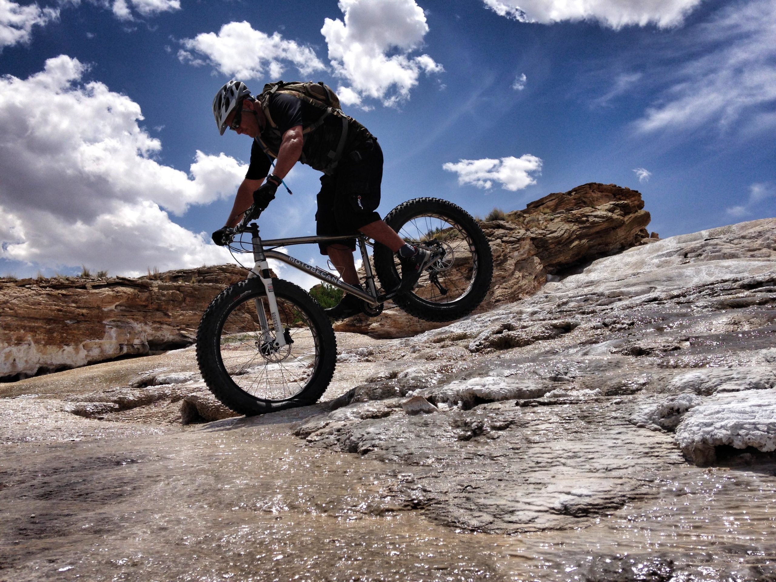 A person riding a mountain bike on a rocky terrain, skillfully balancing as they perform a wheelie. The scene is set under a bright blue sky with white clouds, and the ground appears wet, reflecting light. The surroundings include rocky outcrops and hints of vegetation in the background. White Ridge Bike Trails mountain bike trail.