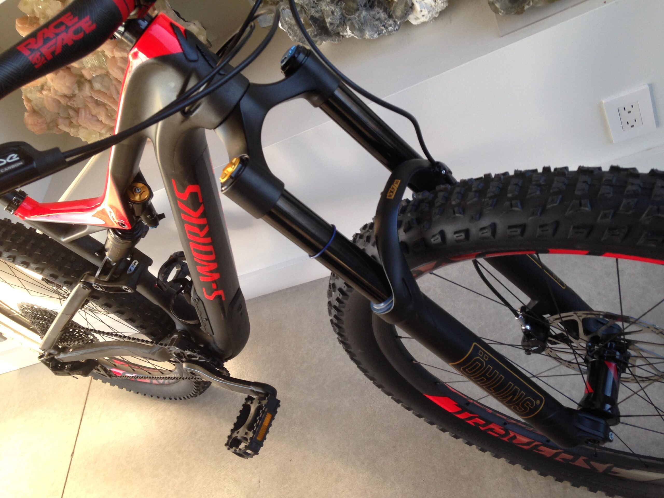 Specialized Stumpjumper FSR 6Fattie: A close-up view of a high-performance mountain bike featuring a sleek black and red frame with the brand name "S-WORKS" prominently displayed. The bike is equipped with thick, knobby tires designed for off-road terrain, and details of the suspension system and pedals are visible, highlighting its advanced engineering. The background is a neutral-colored wall.