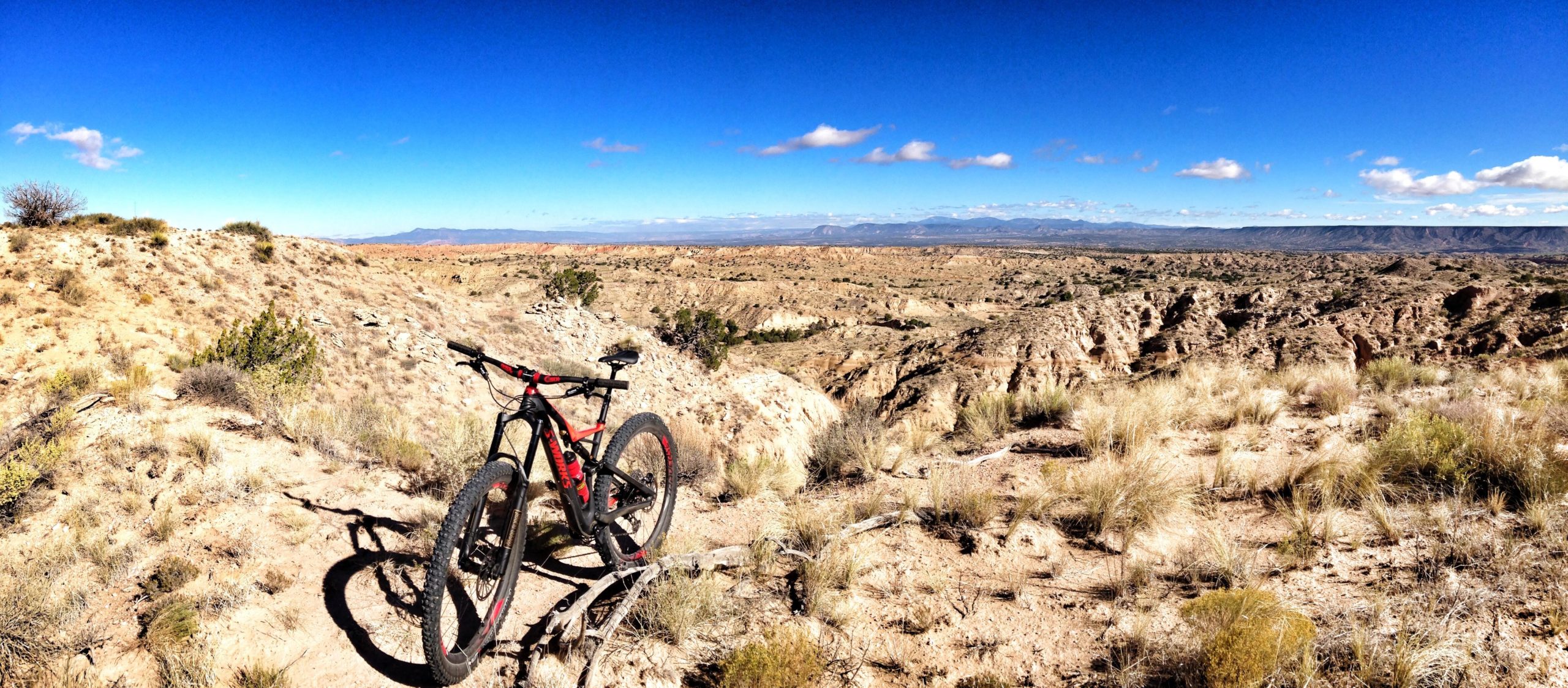 A mountain bike resting on rocky terrain with a vast, arid landscape in the background. The scene features a clear blue sky with a few fluffy clouds and distant mountains, set against dry shrubs and grasses. The composition showcases the beauty of outdoor cycling in a rugged wilderness area. Mariposa Fat Bike Trails mountain bike trail.
