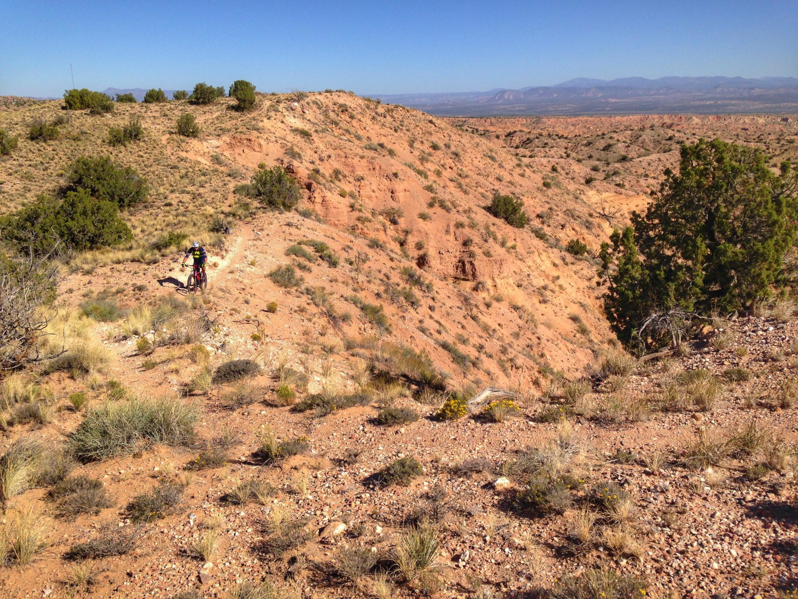 A mountain biker navigating a rocky trail on a hillside with sparse vegetation, surrounded by scenic desert landscape under a clear blue sky. Mariposa Fat Bike Trails mountain bike trail.