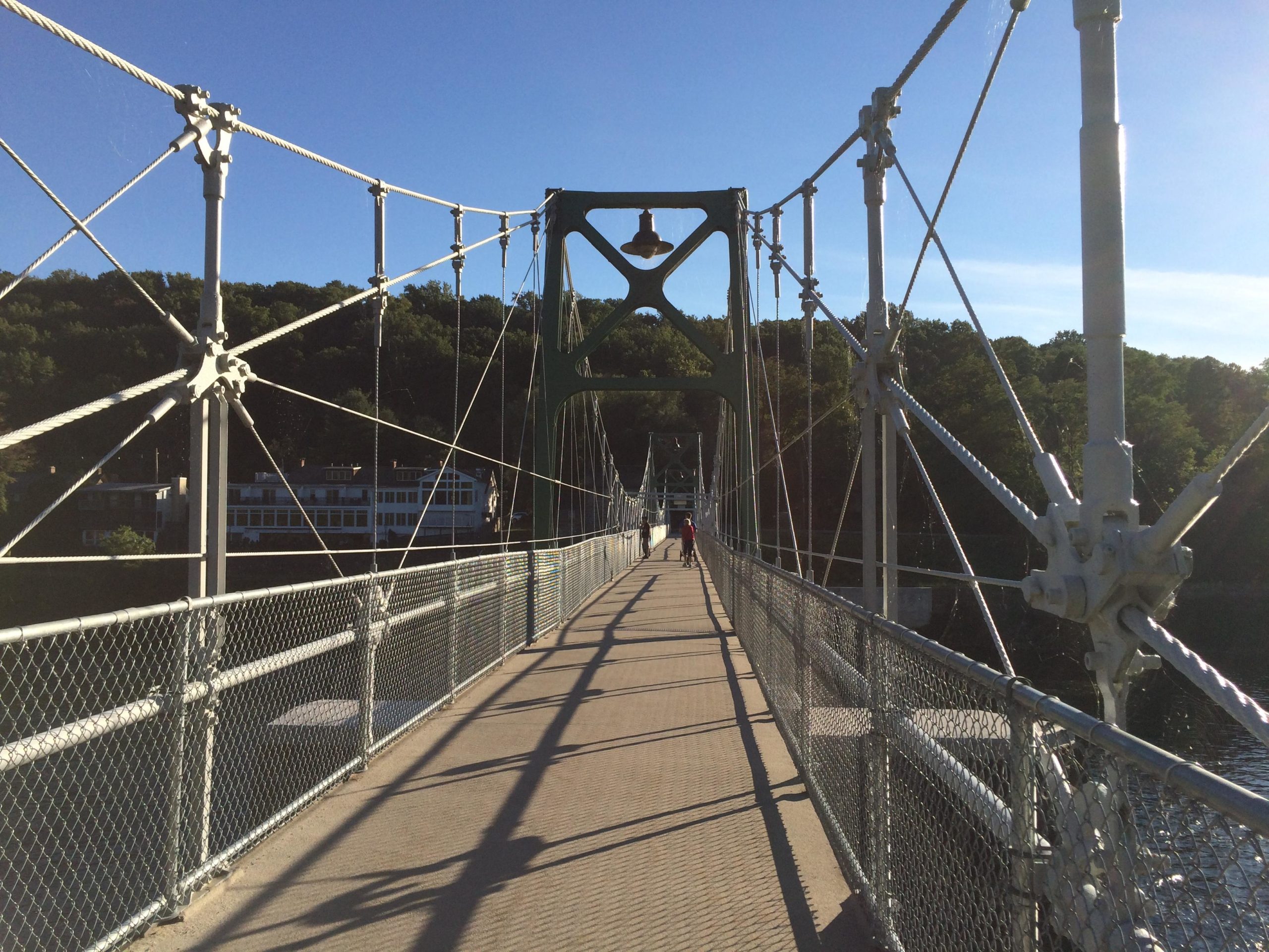Alt tag: "Suspension bridge with a pathway, featuring green metal supports and cable. A person walks towards the camera, with trees and a building visible in the background under a clear blue sky." Bridge To Bridge - D&r Canal mountain bike trail.