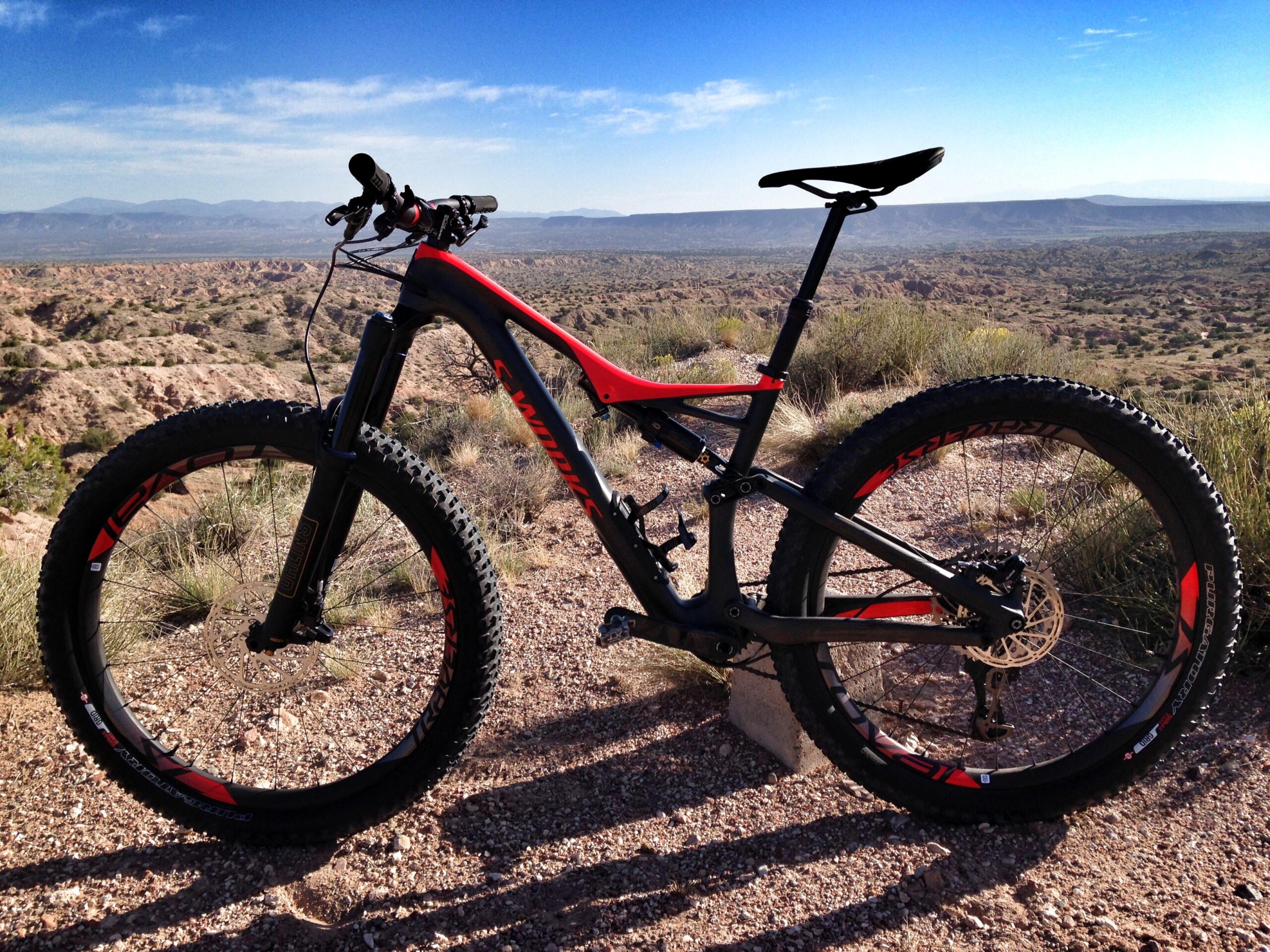 Specialized Stumpjumper FSR 6Fattie: A mountain bike with a black and red frame is parked on rocky terrain, overlooking a vast, scenic landscape with rolling hills under a clear blue sky. The bike features thick tires and a lightweight design, ideal for off-road trails.