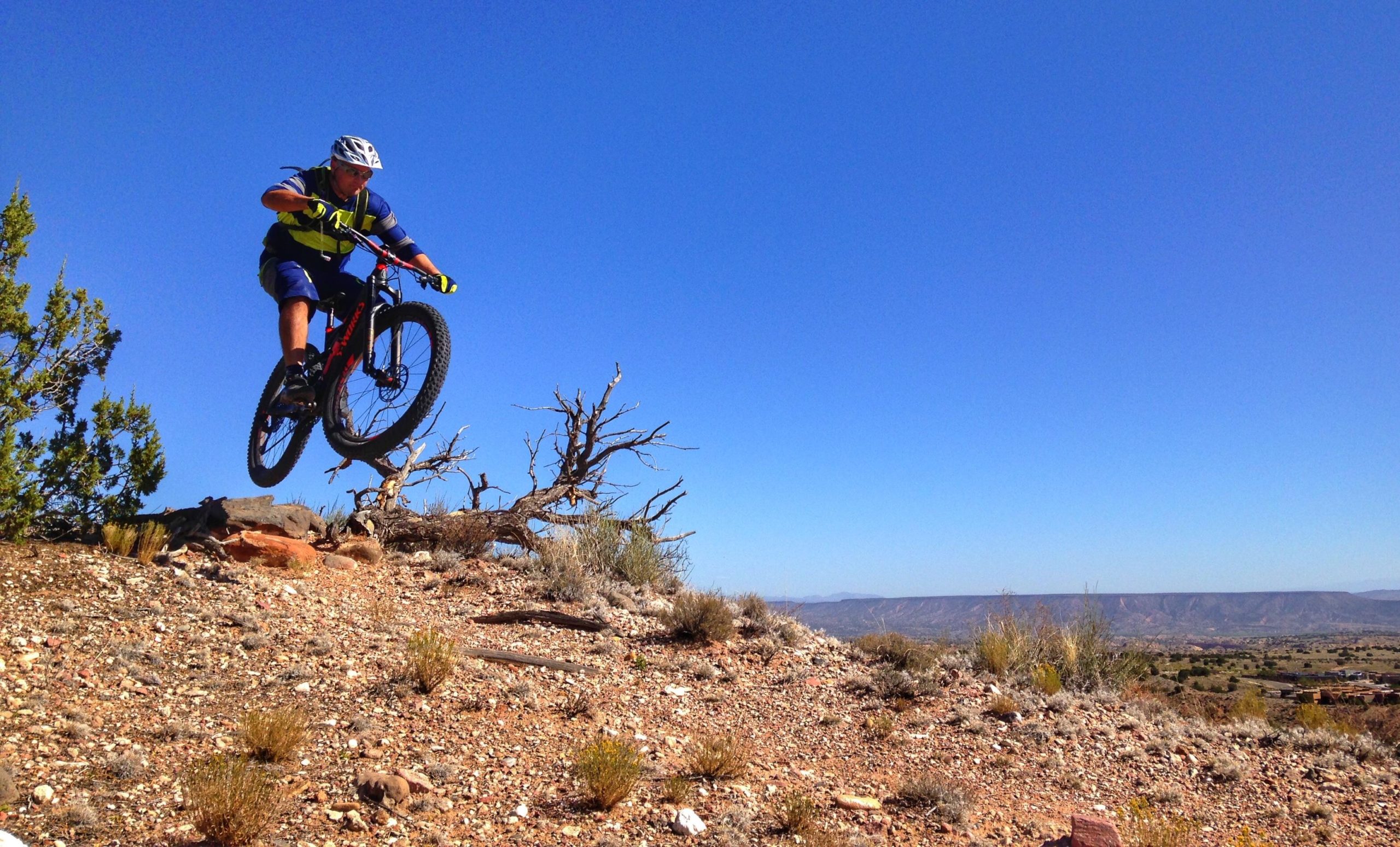 A mountain biker performing a jump over rocky terrain against a clear blue sky, with desert vegetation and distant mountains in the background. Mariposa Fat Bike Trails mountain bike trail.