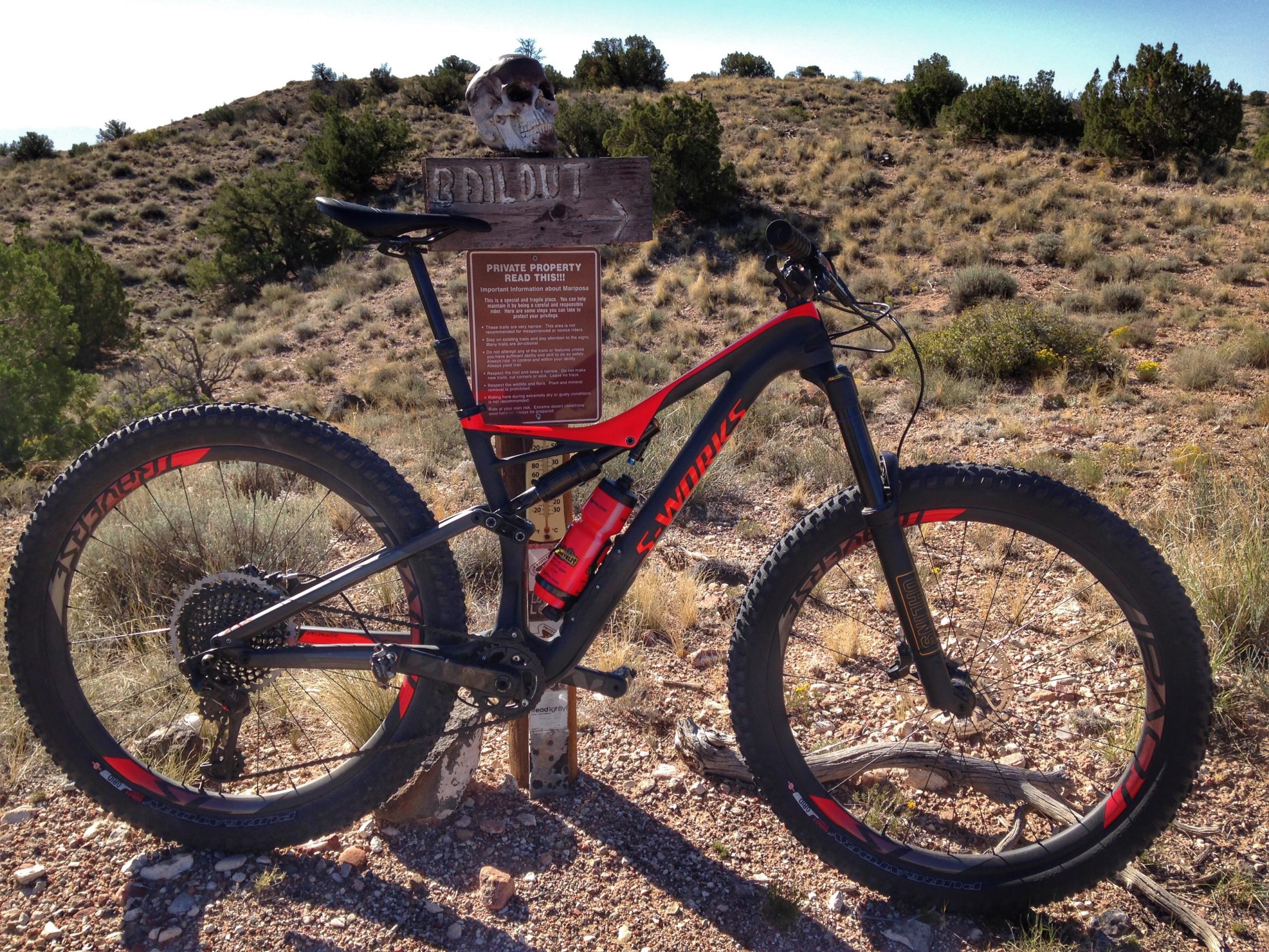 A mountain bike leaning against a wooden signpost in a rural landscape. The sign, titled "Bailout," provides important information and has a small skull attached to it. The terrain is dry, with sparse vegetation and rolling hills in the background under a clear blue sky. Mariposa Fat Bike Trails mountain bike trail.