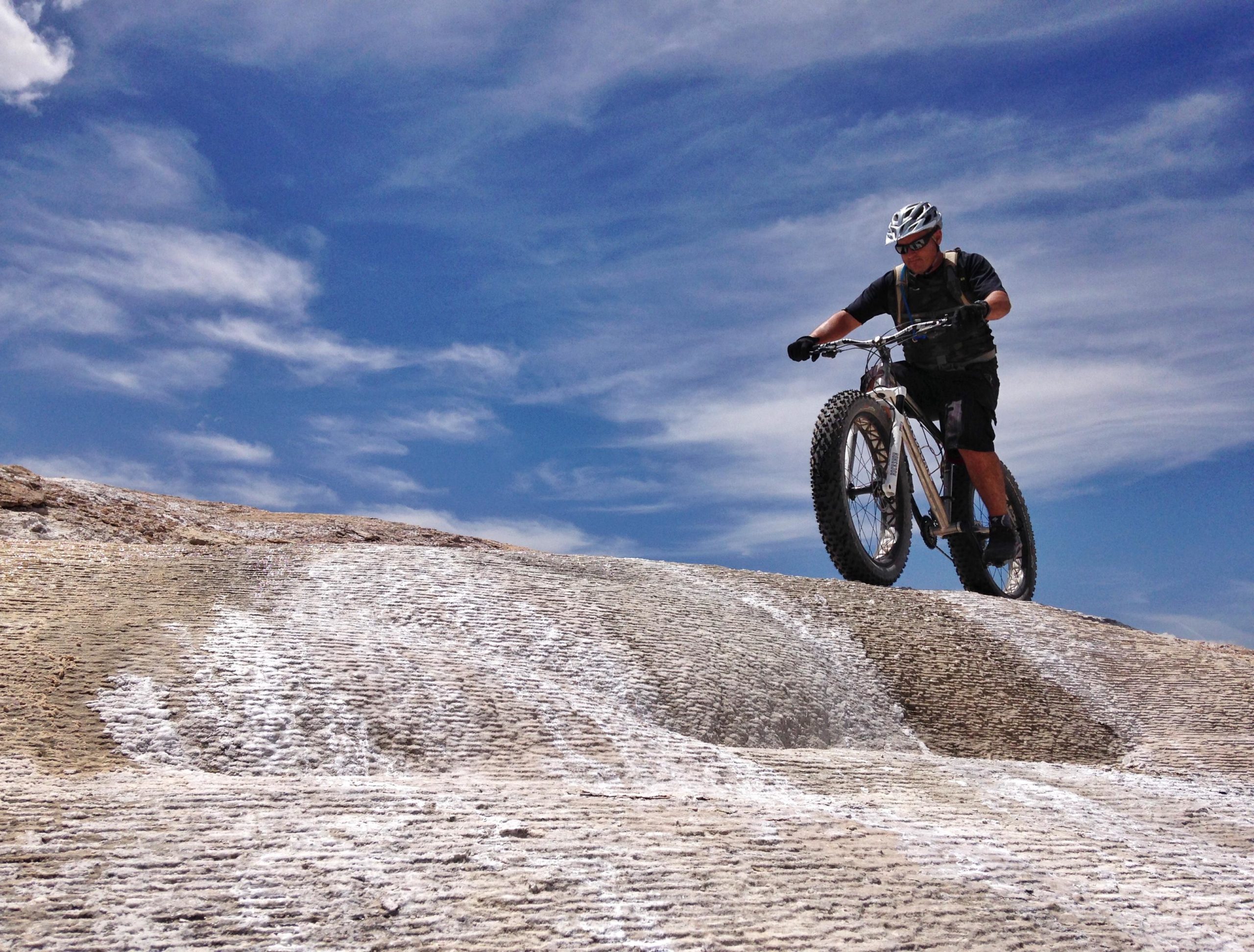 A cyclist riding a fat bike on a rocky terrain under a bright blue sky with scattered clouds. The bike features wide tires designed for uneven surfaces, and the cyclist is focused as they navigate the landscape. White Ridge Bike Trails mountain bike trail.