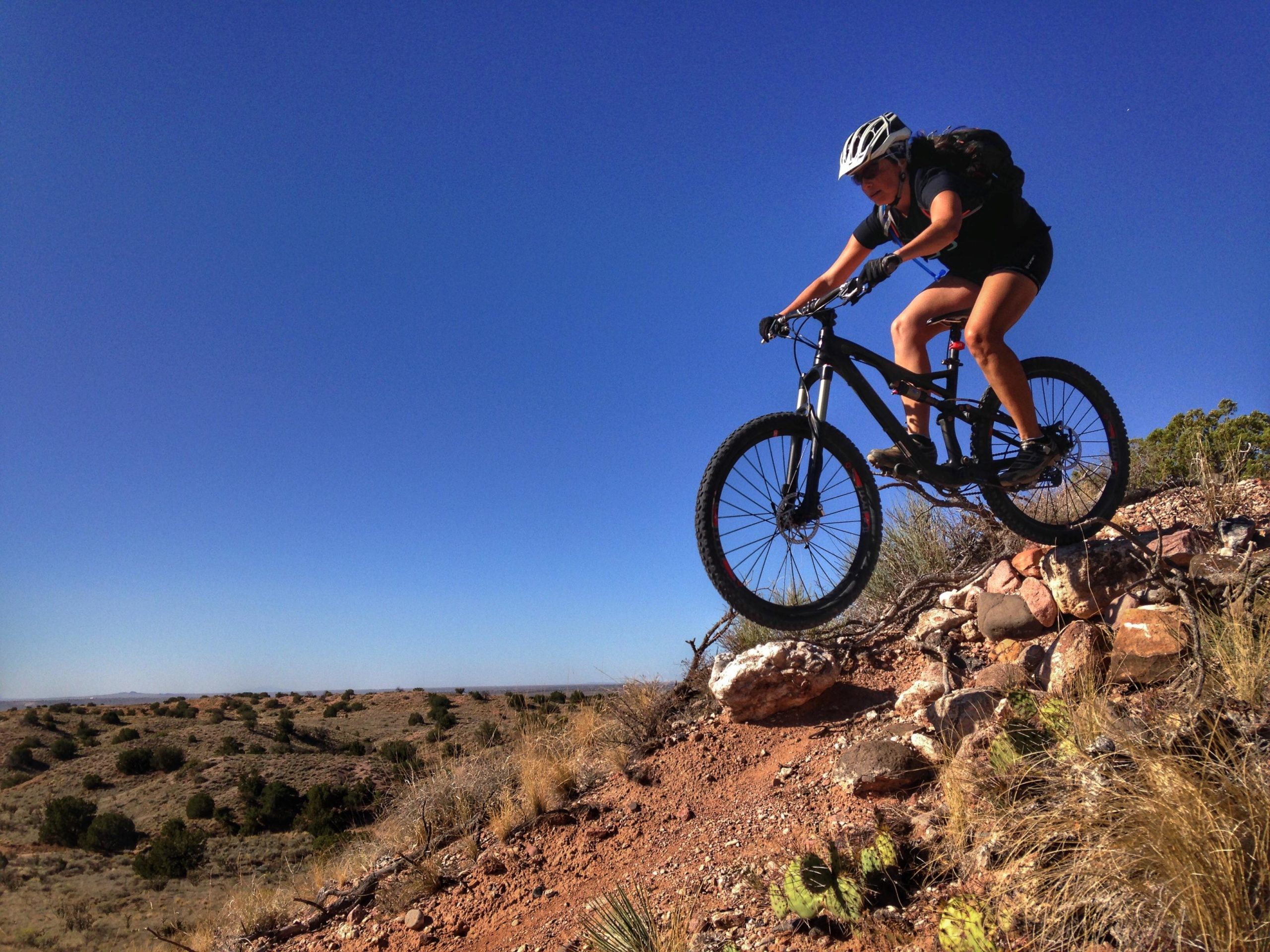 A female mountain biker in a black cycling outfit and helmet jumps off a rocky ledge, with a vast desert landscape and clear blue sky in the background. Mariposa Fat Bike Trails mountain bike trail.
