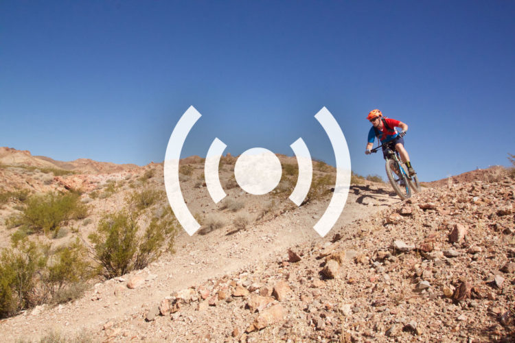 A mountain biker riding down a rocky trail in a rugged desert landscape under a clear blue sky.