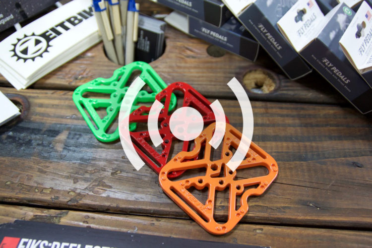 Three colorful bike pedals in green, red, and orange are displayed on a wooden surface, surrounded by marketing materials and product boxes.
