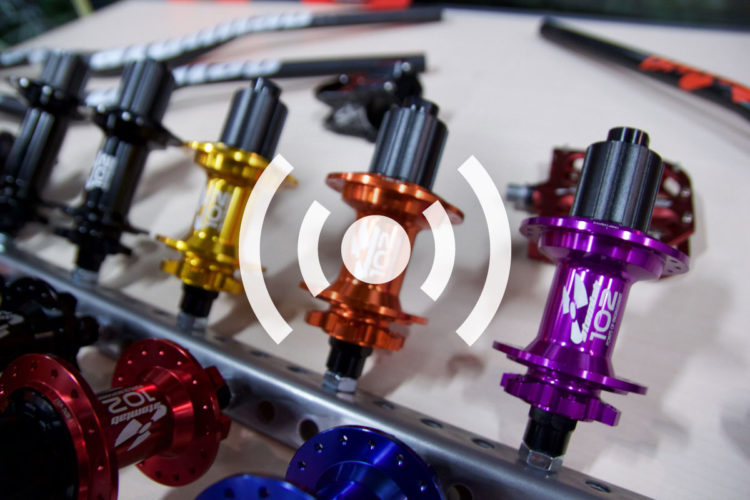 A close-up image of colorful bicycle hubs arranged on a display. The hubs are in various colors, including black, gold, orange, red, and purple, highlighting their detailed designs and branding. A graphic symbol representing connectivity is overlaid in the center.