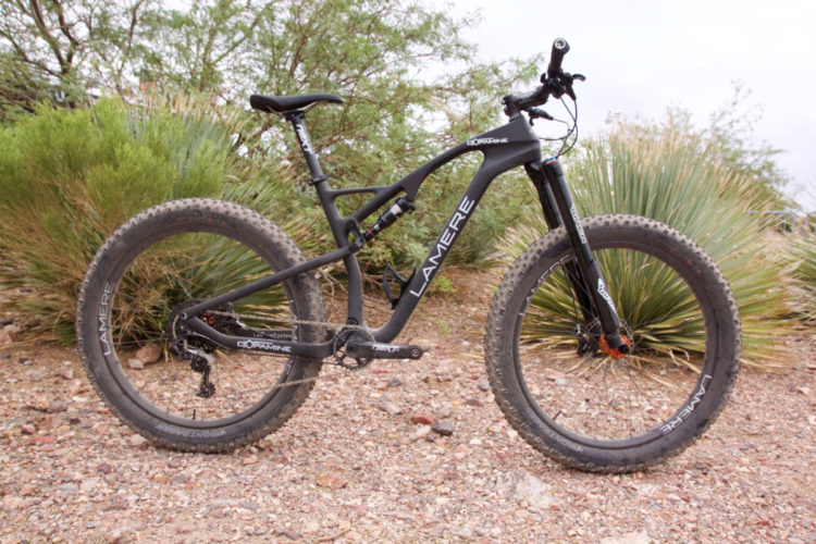 A black mountain bike with wide tires, positioned in a rocky desert environment surrounded by shrubs and vegetation. The bike features a sleek frame with the brand name "Lamere" visible. The image captures the bike at an angle that highlights its design and tires, set against a natural backdrop.