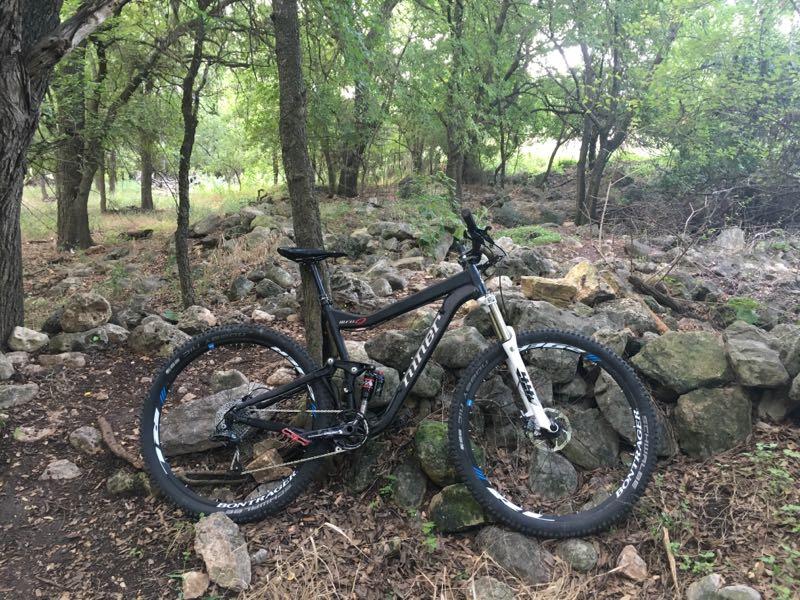 A black mountain bike resting against a rocky wall in a wooded area, surrounded by trees and greenery. The bike features large tires and is designed for off-road trails. Salado Creek mountain bike trail.