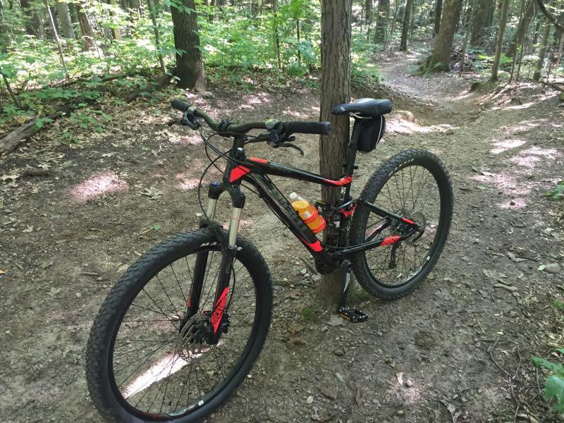 A black and red mountain bike resting against a tree on a dirt trail in a forested area, with greenery surrounding the path. The bike has a water bottle attached to the frame and trails leading off into the dense woods. Pontiac Lake mountain bike trail.