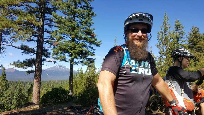A bearded man wearing a mountain biking helmet and a black shirt with blue accents stands in a forested area, smiling at the camera. In the background, tall trees and a mountain landscape are visible under a clear blue sky. Dinah Moe Humm mountain bike trail.