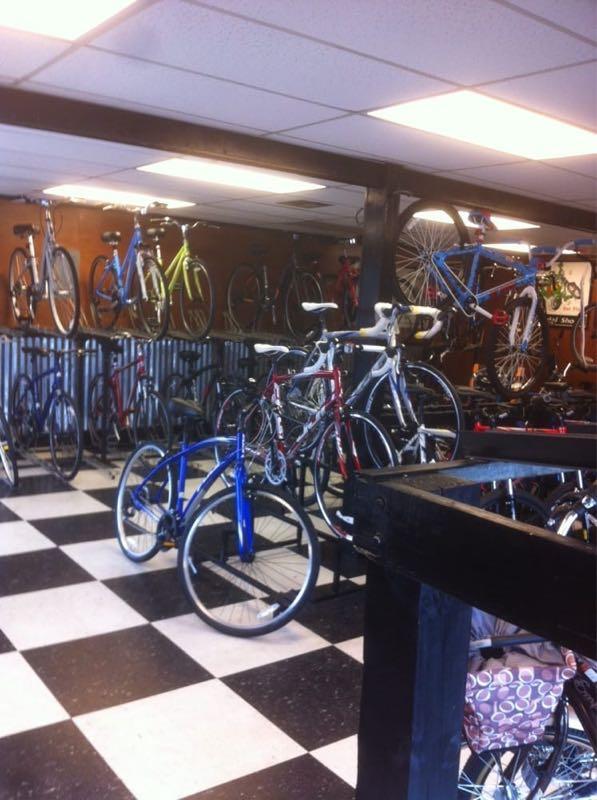 Interior of a bike shop featuring a variety of bicycles displayed on walls and racks, with a checkered black and white floor. The shop is well-lit, showcasing bicycles in different colors, including blue, red, green, and white.