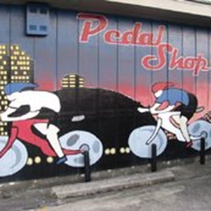 A colorful mural depicting two cartoon cyclists racing, with a city skyline in the background. The mural features the text "Pedal Shop" in bold letters, and the scene is set against a gradient sunset.