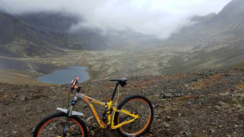 A yellow mountain bike positioned on rocky terrain, overlooking a vast valley with a small lake, surrounded by misty mountains under a cloudy sky. Power Line Pass To Indian mountain bike trail.