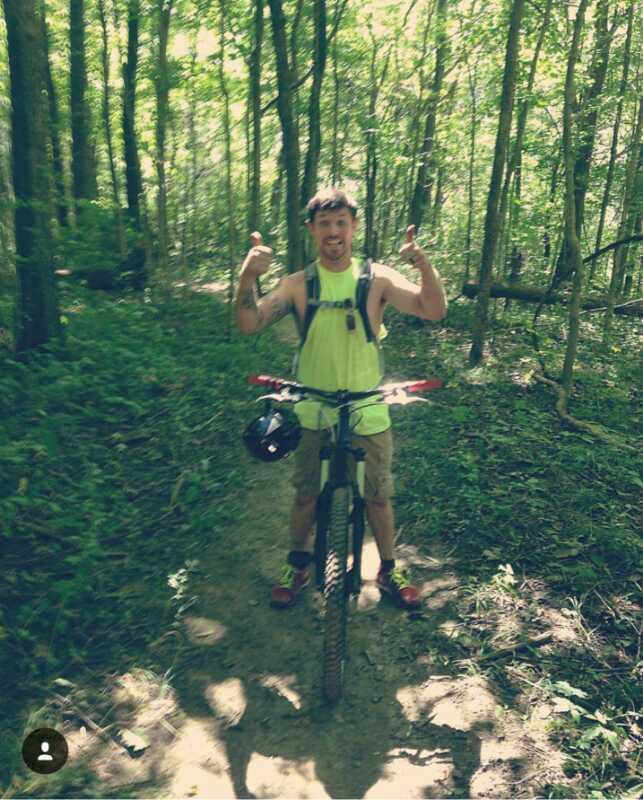 A person wearing a bright yellow tank top and shorts stands on a mountain bike, giving a thumbs-up gesture. They are surrounded by a lush, green forest, and the sunlight filters through the trees, illuminating the scene. The path is dirt and surrounded by foliage. Hueston Woods State Park mountain bike trail.