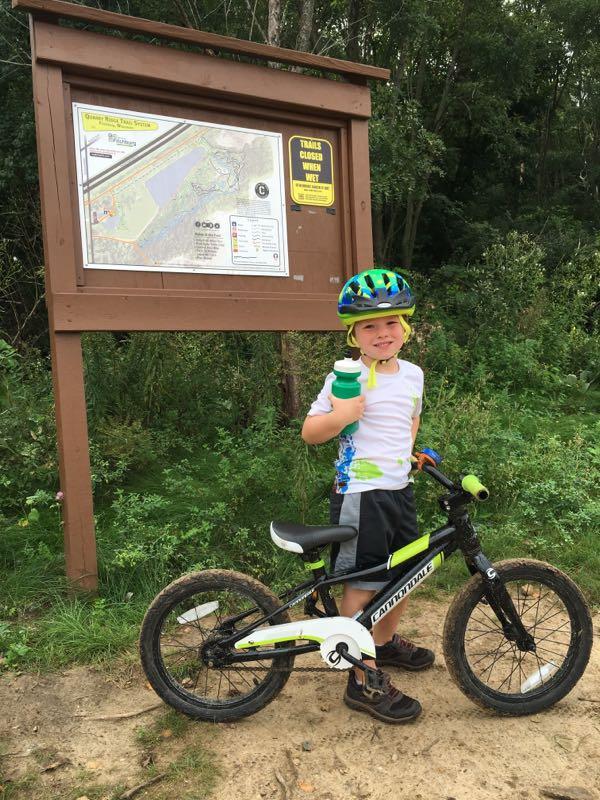 A smiling child wearing a blue and white helmet and a colorful shirt stands beside a bicycle. The child is holding a green water bottle, and a trail map sign is visible in the background, surrounded by trees and greenery. The scene is set on a dirt path, suggesting an outdoor recreational area. Quarry Ridge Park mountain bike trail.